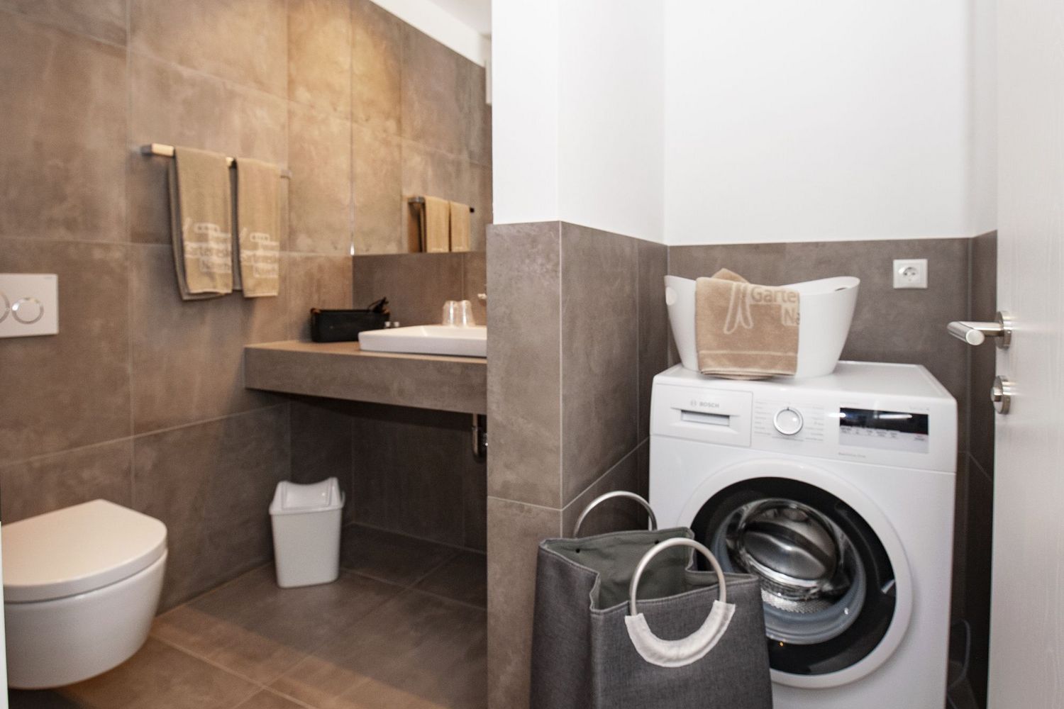 Ein Badezimmer mit modernen braunen Fliesen, Toilette, Waschbecken und einer Waschmaschine mit Wäschekorb.