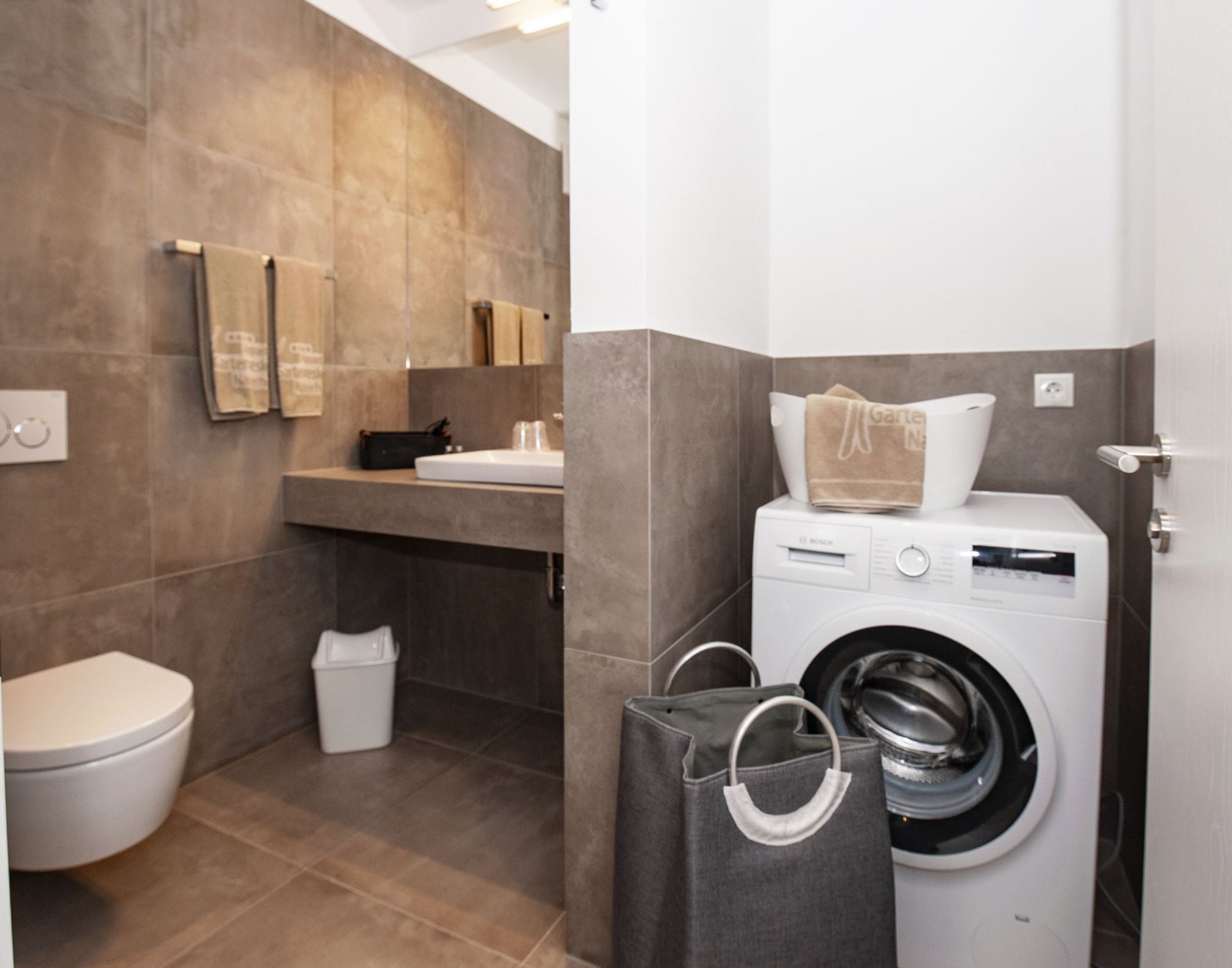 Ein Badezimmer mit modernen braunen Fliesen, Toilette, Waschbecken und einer Waschmaschine mit Wäschekorb.
