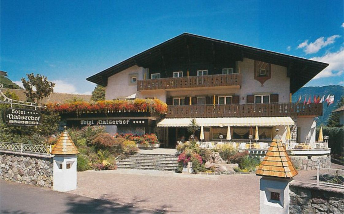 Ein traditionelles vierstöckiges Hotelgebäude im alpinen Stil mit Holzbalkonen, blumenbesetzter Terrasse und einer Treppe zum Eingang vor einem strahlend blauen Himmel.