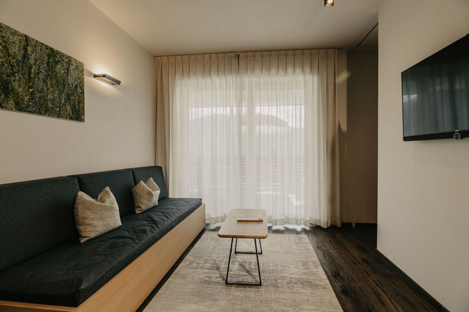 Ein modern eingerichtetes Wohnzimmer mit einem dunkelgrauen Sofa, einem kleinen Holztisch, einem Teppich sowie einem Fernseher und Vorhängen vor einer Glasfront.