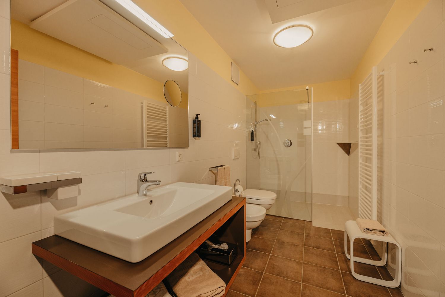 Ein modernes Badezimmer mit einem Waschbecken auf einem Holzunterschrank, einer ebenerdigen Dusche, einer Toilette, einem Bidet und braunen Fliesen.