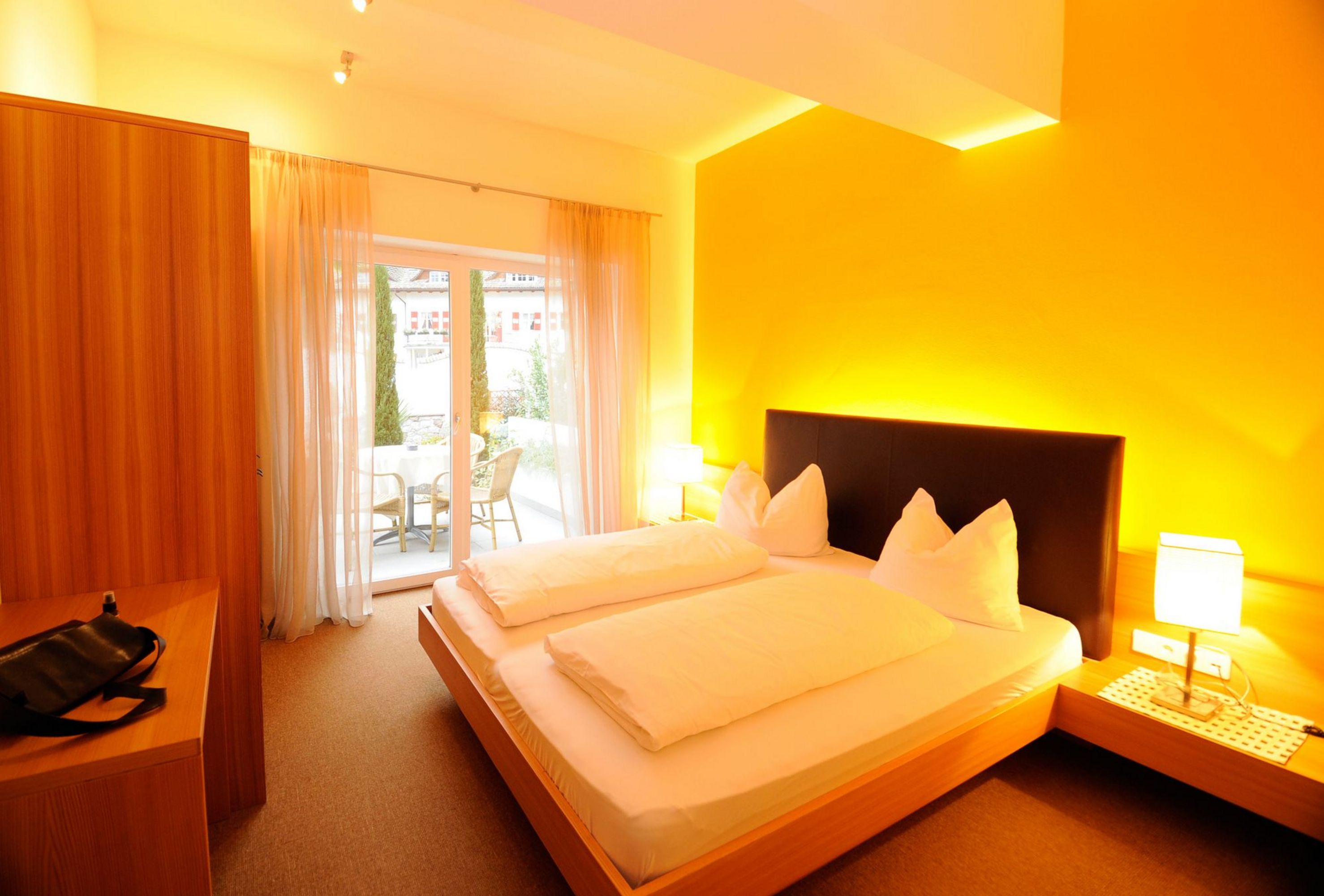 Ein gemütliches Hotelzimmer mit einem Doppelbett, warmen gelben Wandfarben und einer Terrassentür, die nach draußen führt.