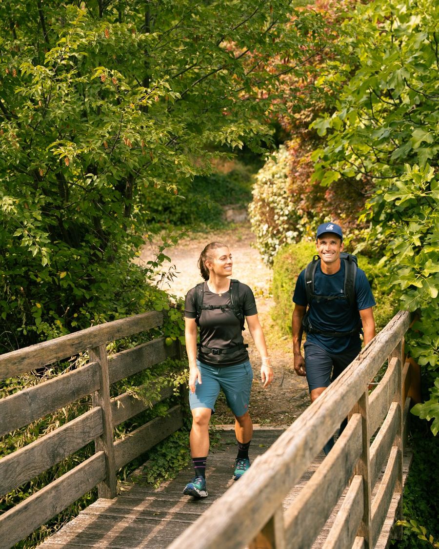 Ein Wanderpaar mit Rucksäcken überquert lächelnd eine kleine Holzbrücke in einer grünen, waldreichen Umgebung.