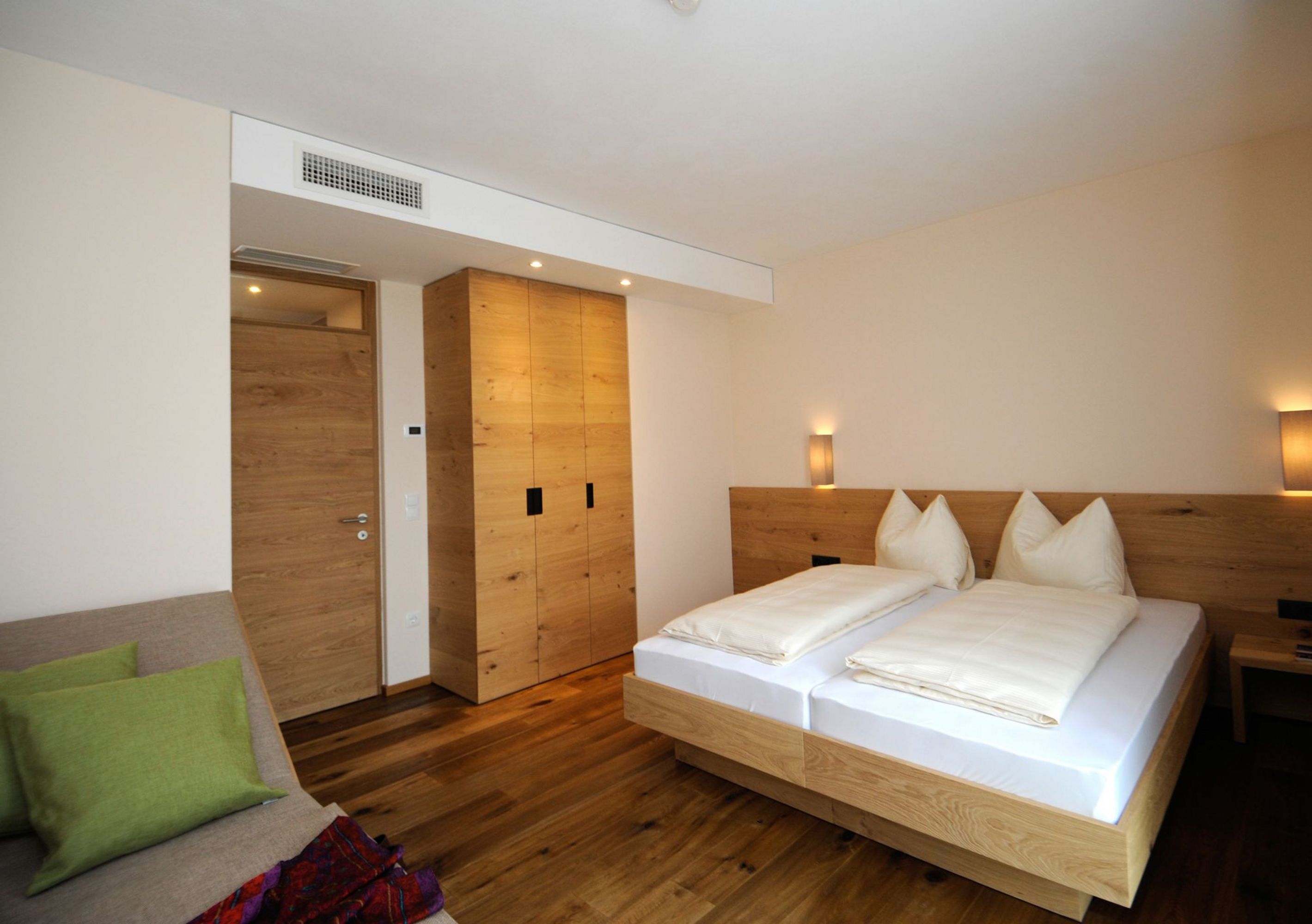 Ein modernes Hotelzimmer mit Zirbenholzmöbeln, einem Doppelbett, einem Kleiderschrank und einer kleinen Sitzecke mit grünem Kissen.