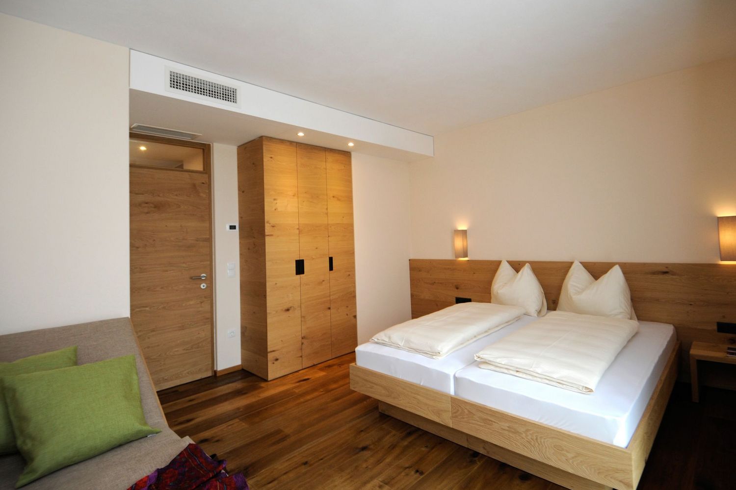 Ein modernes Hotelzimmer mit Zirbenholzmöbeln, einem Doppelbett, einem Kleiderschrank und einer kleinen Sitzecke mit grünem Kissen.