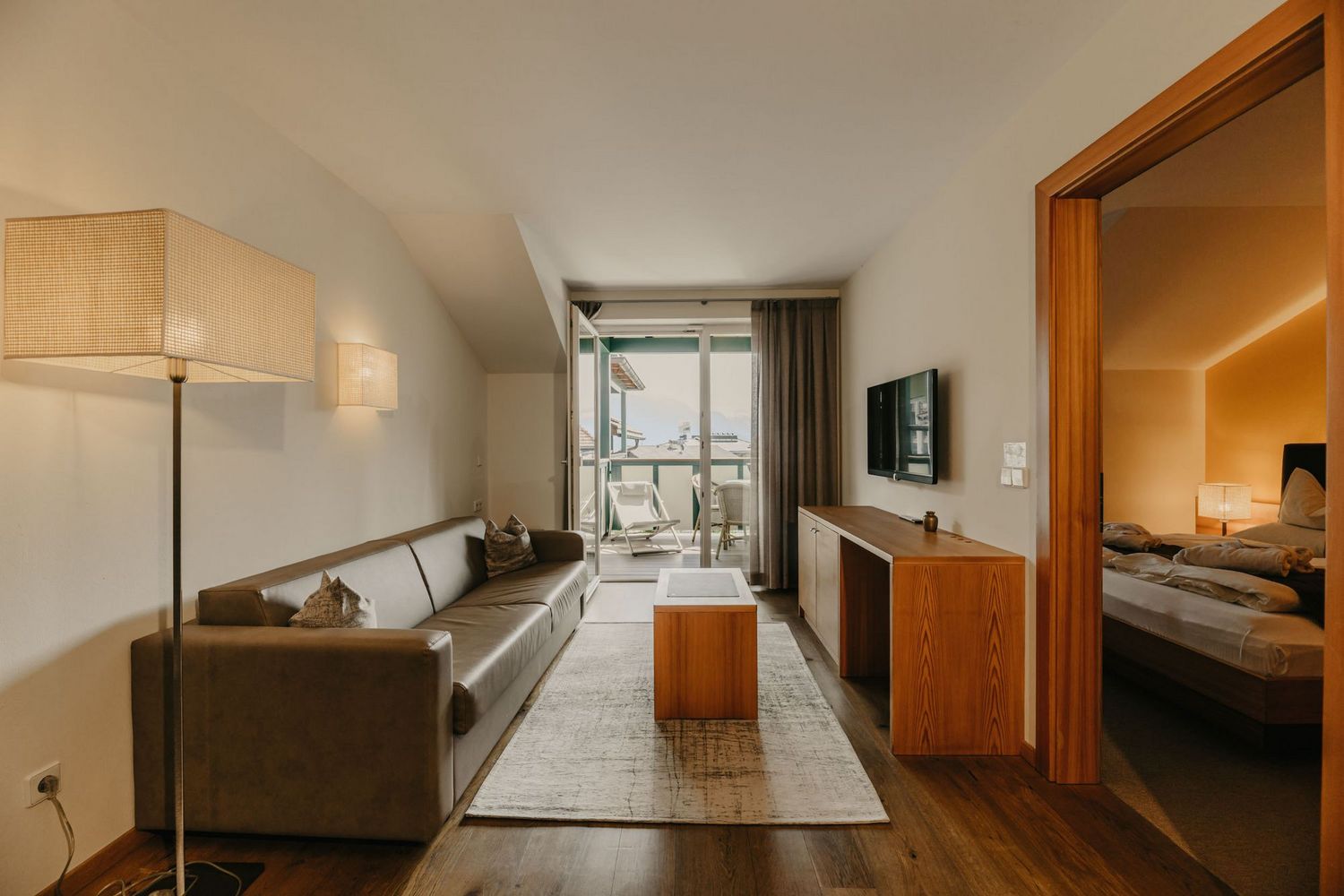 Ein einladendes Hotelzimmer mit braunem Ledersofa, einem Couchtisch auf einem Teppich, einem TV-Board und Blick auf einen Balkon, angrenzend ist ein weiteres Schlafzimmer durch einen Türrahmen zu sehen.