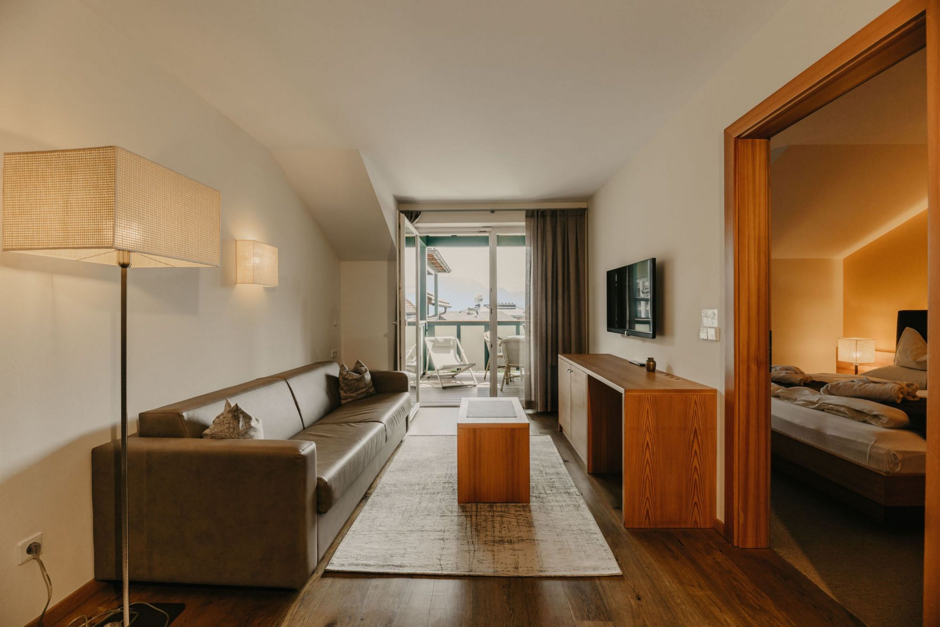 Ein einladendes Hotelzimmer mit braunem Ledersofa, einem Couchtisch auf einem Teppich, einem TV-Board und Blick auf einen Balkon, angrenzend ist ein weiteres Schlafzimmer durch einen Türrahmen zu sehen.