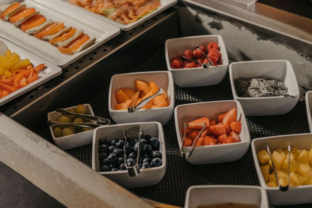 Ein Buffet mit mehreren eckigen weißen Schalen, die mit verschiedenen frischen Obstsorten wie Erdbeeren, Blaubeeren, Melone und Himbeeren gefüllt sind, sowie Platten mit Tomate-Mozzarella im Hintergrund.