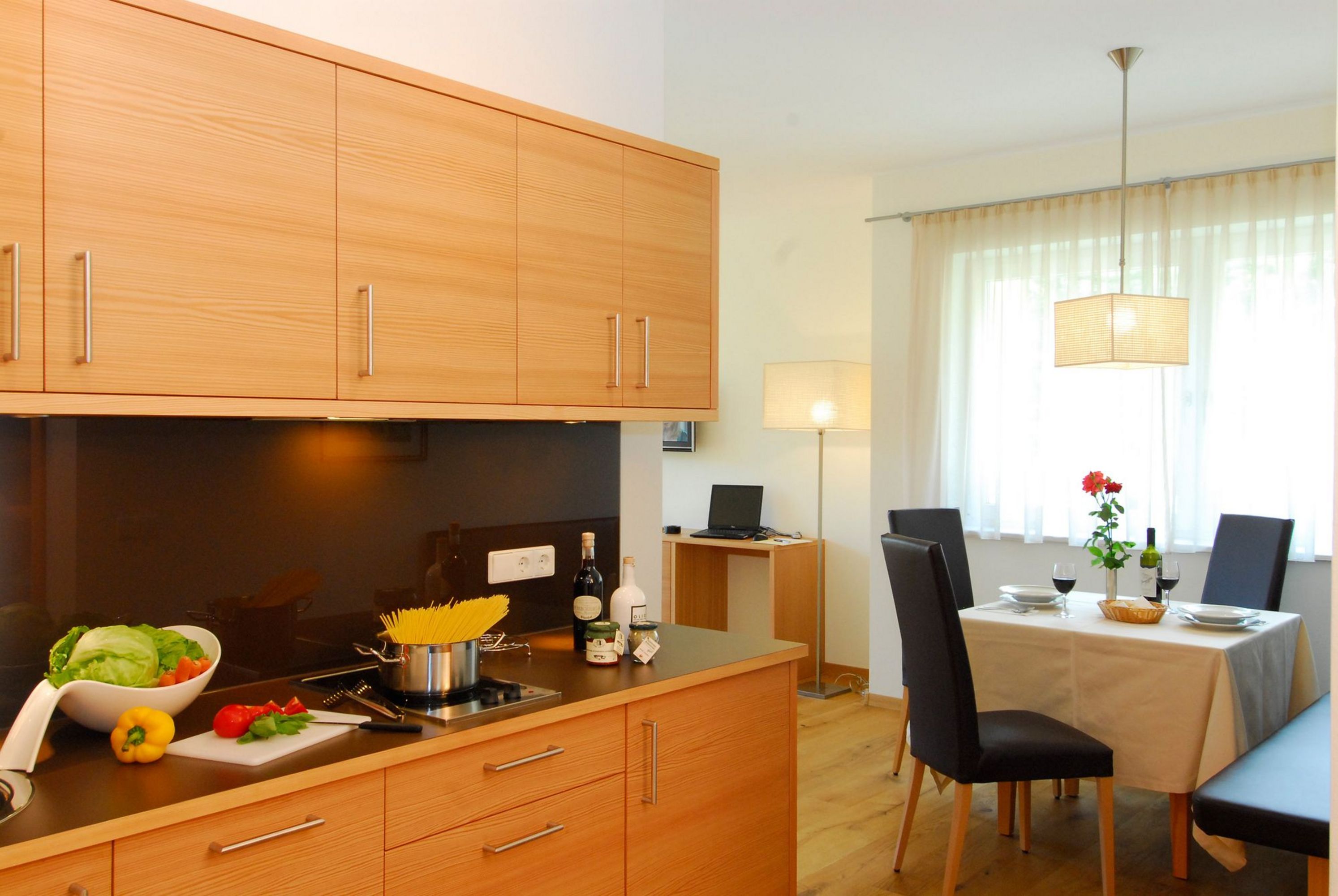 Eine moderne Küche mit hellen Holzfronten und Arbeitsplatte; auf der Arbeitsfläche stehen eine Salatschüssel, eine rote Paprika und ein Topf mit Spaghetti. Im Hintergrund ist ein gedeckter Esstisch mit schwarzen Stühlen zu sehen.