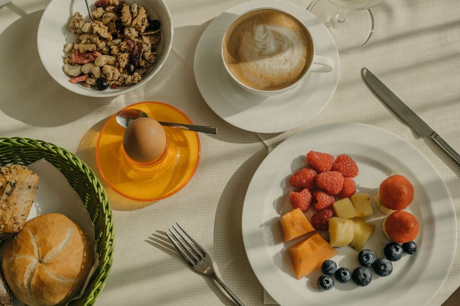 Eine Vogelperspektive auf einen gedeckten Frühstückstisch mit einer Tasse Cappuccino, einer Schüssel Müsli, einem weichgekochten Ei, einem Teller mit frischem Obst und einem Brotkorb.