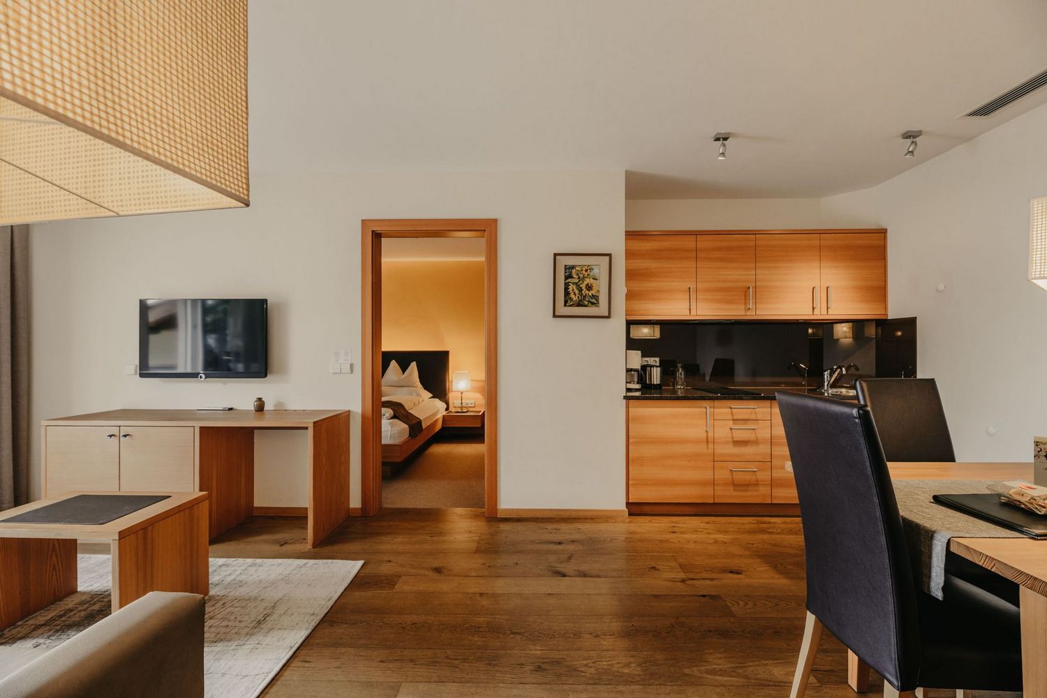 Ein modern eingerichtetes Apartment mit Holzboden, bestehend aus einer offenen Küche mit hellen Holzfronten, einem Essbereich mit dunklen Stühlen und einem Blick durch einen Türrahmen in ein angrenzendes Schlafzimmer mit Doppelbett.