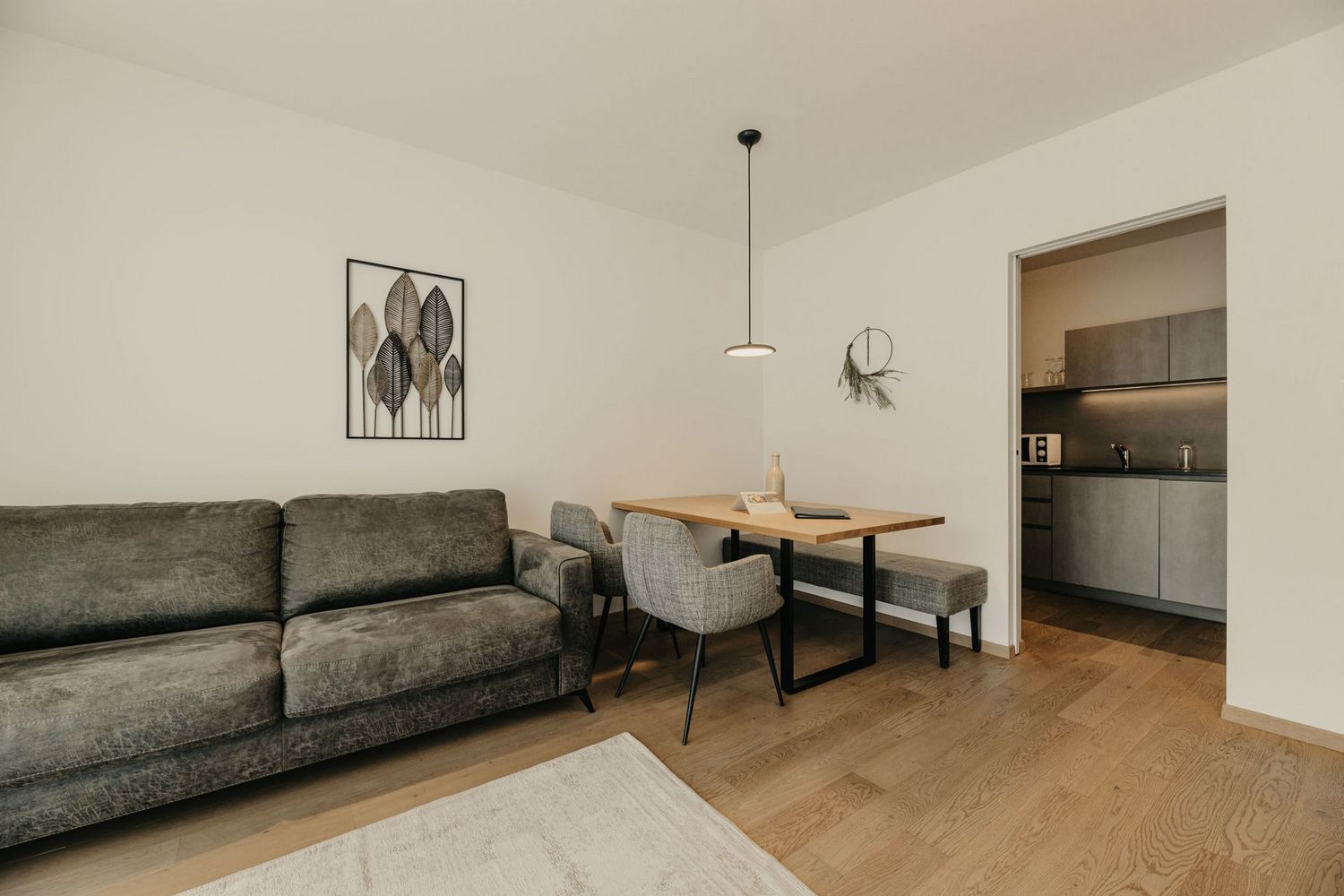 Ein minimalistisch eingerichteter Wohn- und Essbereich mit einem grauen Sofa, einem Holztisch, Stühlen und einer Bank auf hellem Parkettboden, mit Blick in eine angrenzende moderne Küche.