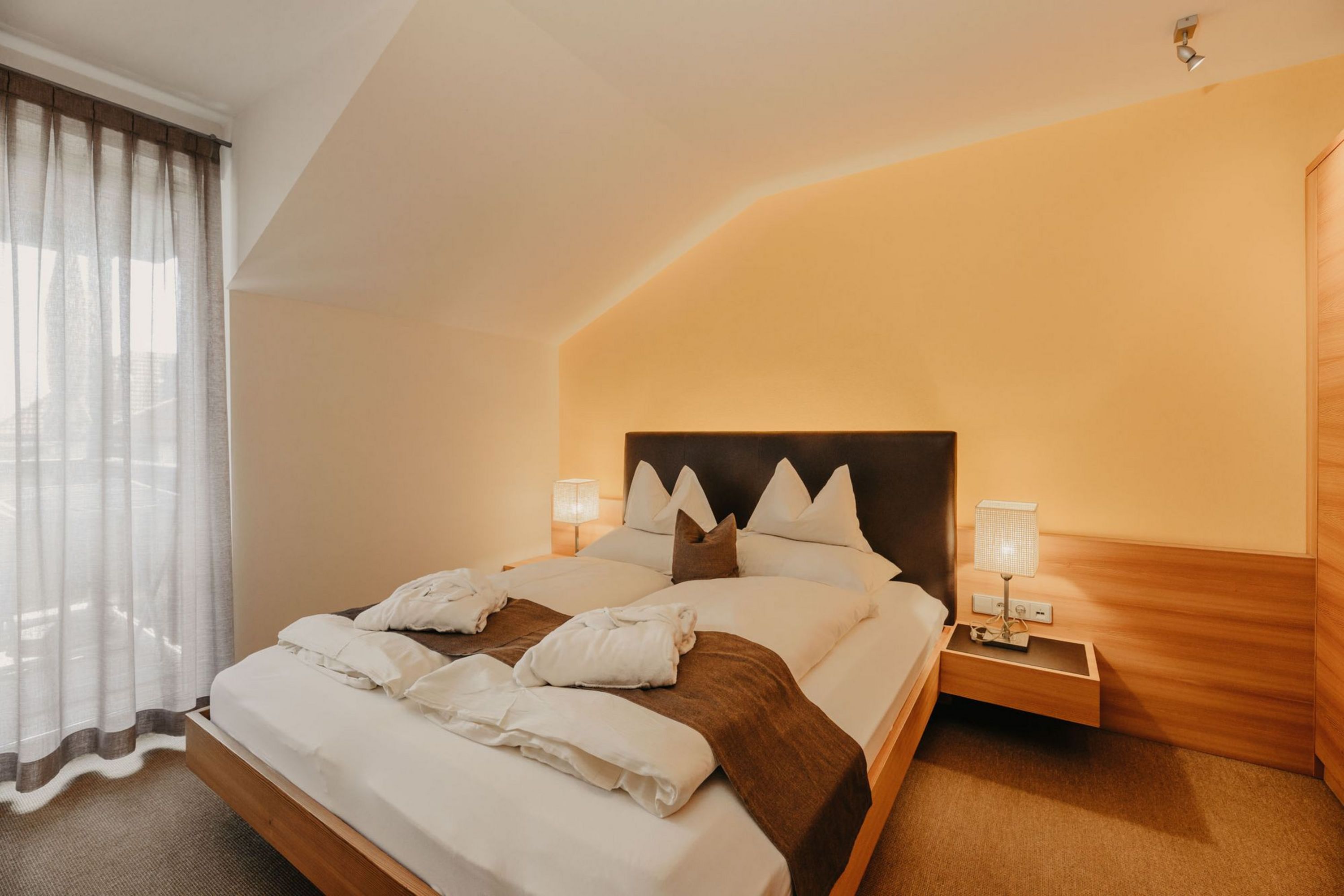 Ein gemütliches Hotelzimmer mit einem Doppelbett, auf dem zwei weiße Bademäntel liegen. Die Wand hinter dem Bett ist gelb gestrichen und es gibt ein Fenster mit einem langen, grauen Vorhang.