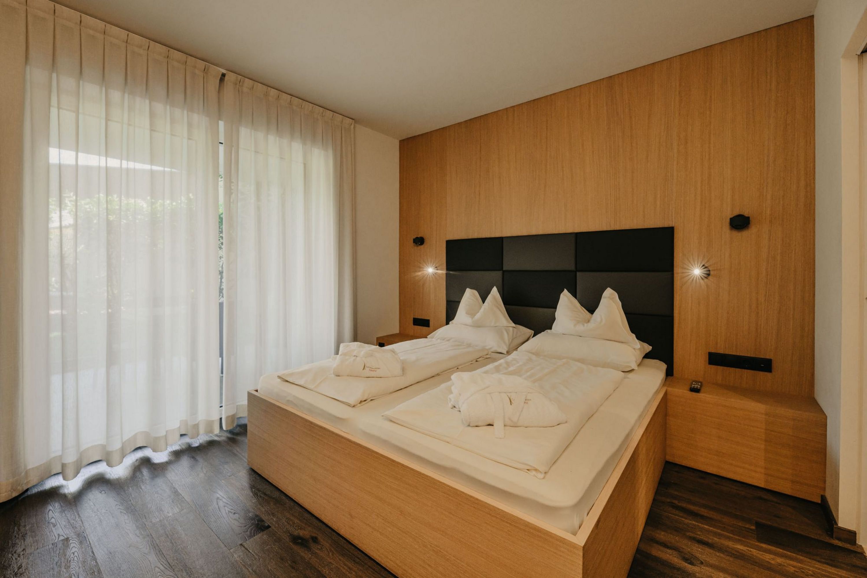 Ein modernes Hotelzimmer mit einem Doppelbett aus hellem Holz, weißen Bettdecken, Bademänteln und einer dunklen gepolsterten Rückwand vor bodentiefen Fenstern mit hellen Vorhängen.