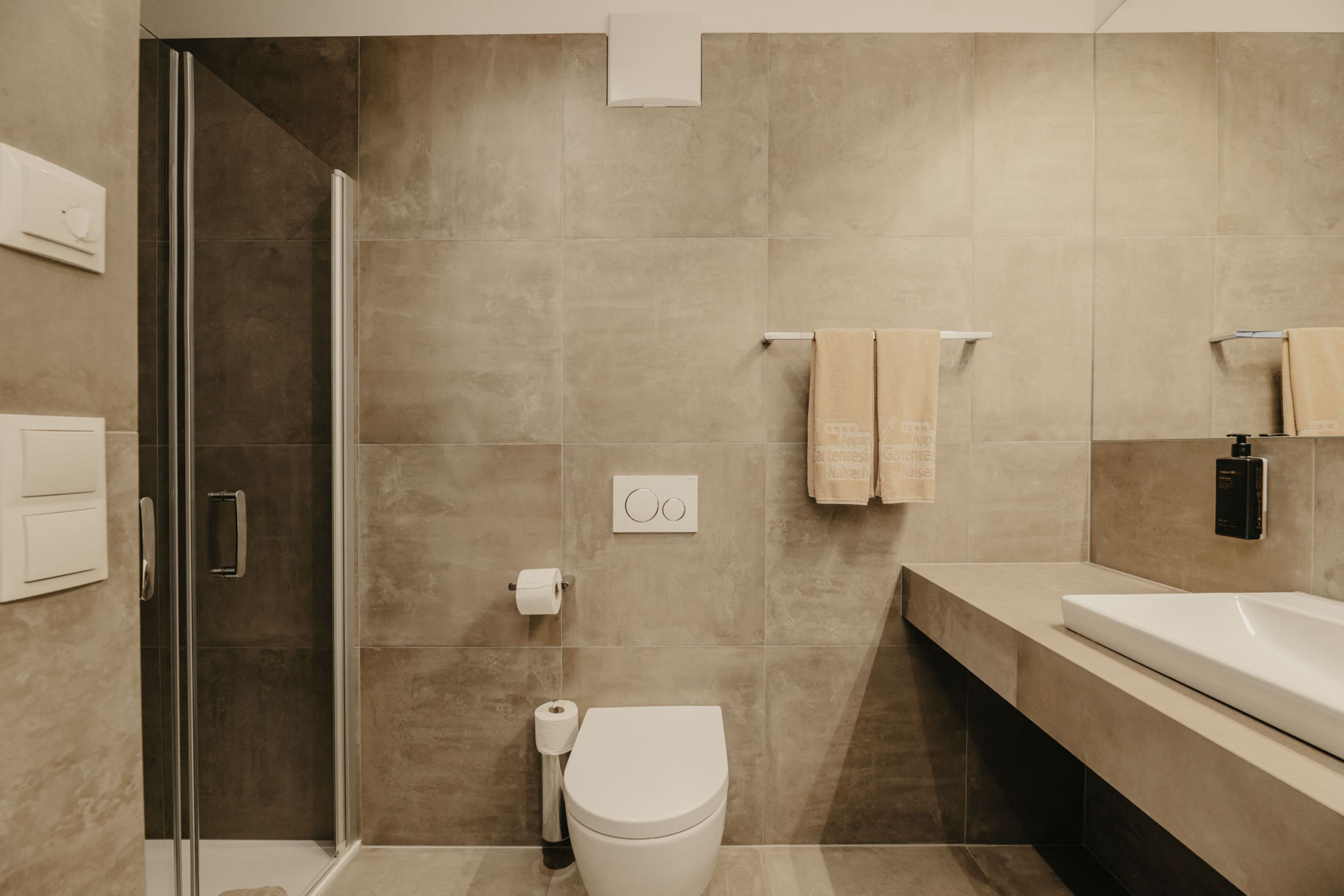 Ein modernes Badezimmer mit grauen Fliesen, bestehend aus einer Glasdusche links, einer hängenden Toilette in der Mitte und einem Waschbecken rechts.