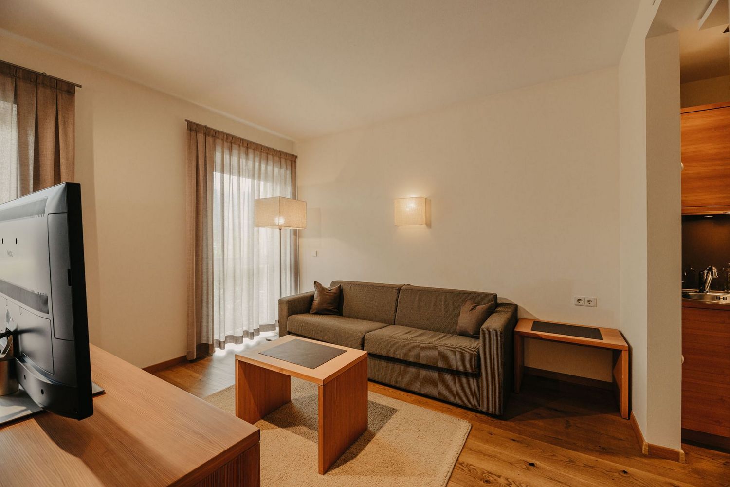 Ein gemütlicher Wohnbereich mit einem grauen Sofa, einem Holztisch, einem Fernseher und einer integrierten Küchenzeile in sanften Brauntönen.