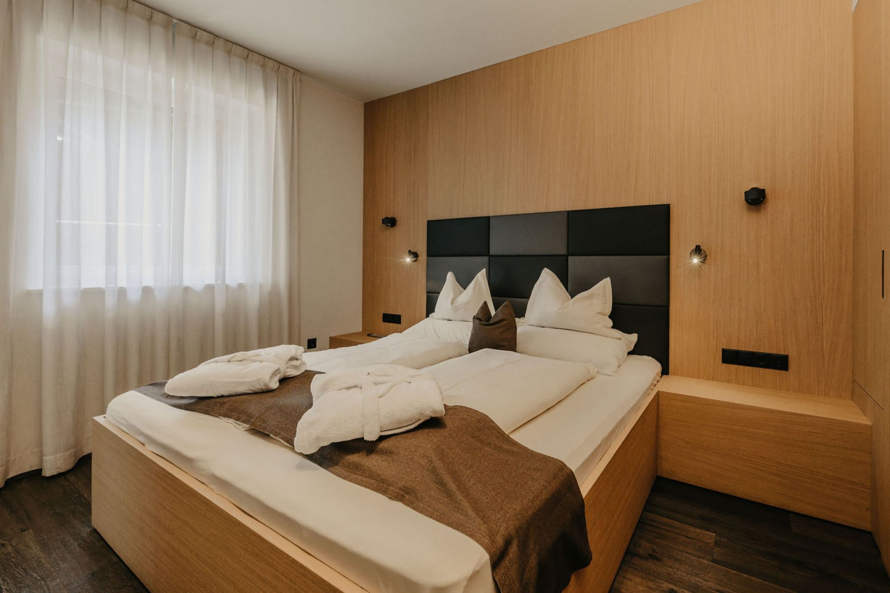 Ein modernes Hotelzimmer mit einem Doppelbett aus hellem Holz, einem dunkelgrauen gepolsterten Kopfteil und zwei weißen Bademänteln auf dem Bett.