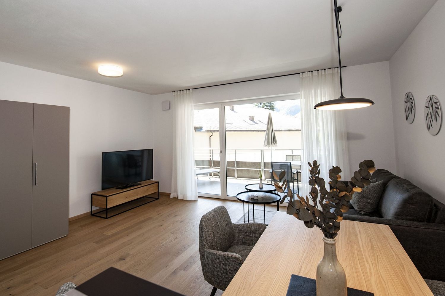 Ein helles Wohnzimmer mit Parkettboden, modernem Sofa, einem Esstisch und einer großen Terrassentür mit Blick nach draußen.