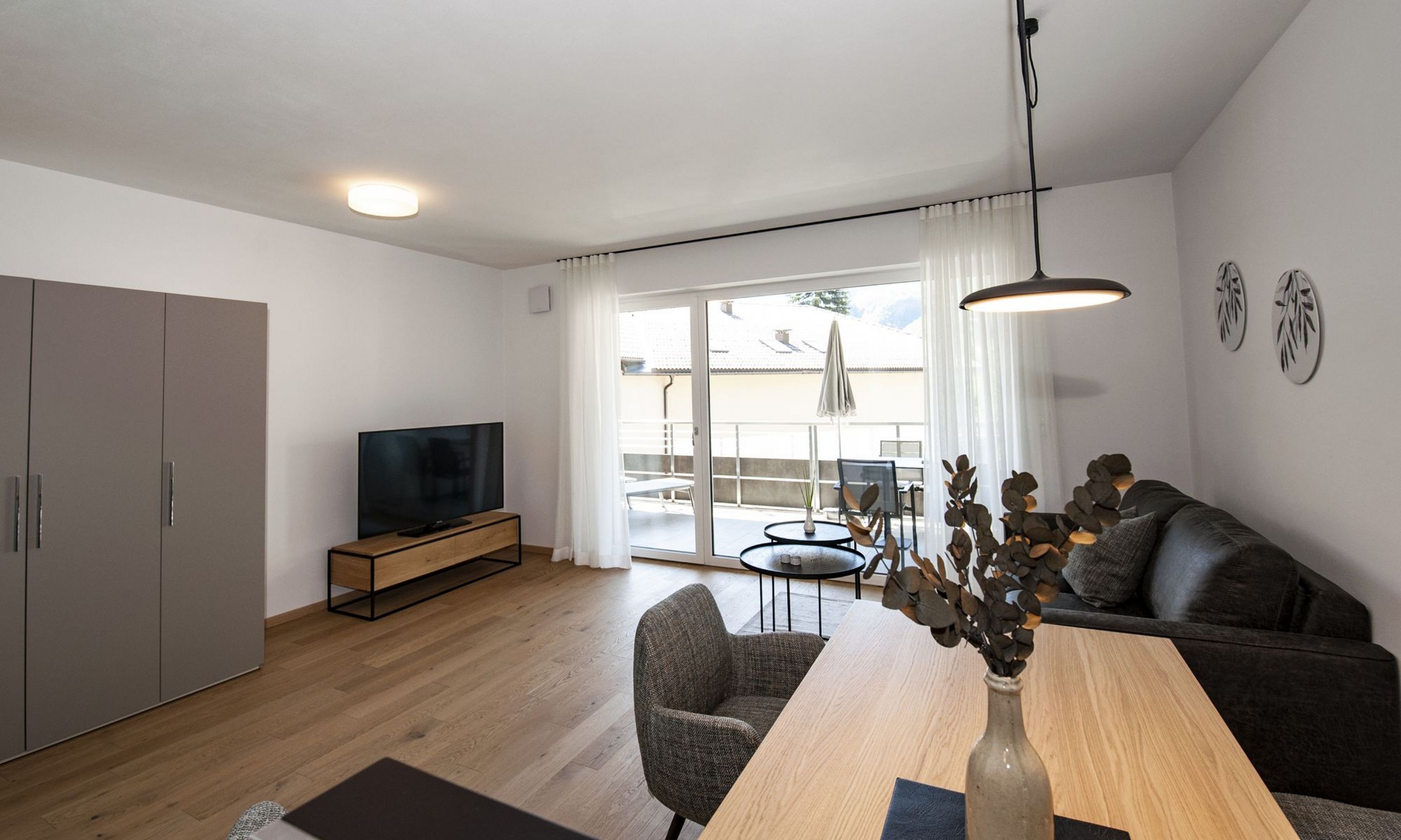 Ein helles Wohnzimmer mit Parkettboden, modernem Sofa, einem Esstisch und einer großen Terrassentür mit Blick nach draußen.