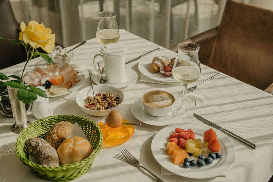 Ein reich gedeckter Frühstückstisch mit Brot, Obst, Müsli, Aufschnitt, Kaffee und Sekt unter sonnigem Lichteinfall.