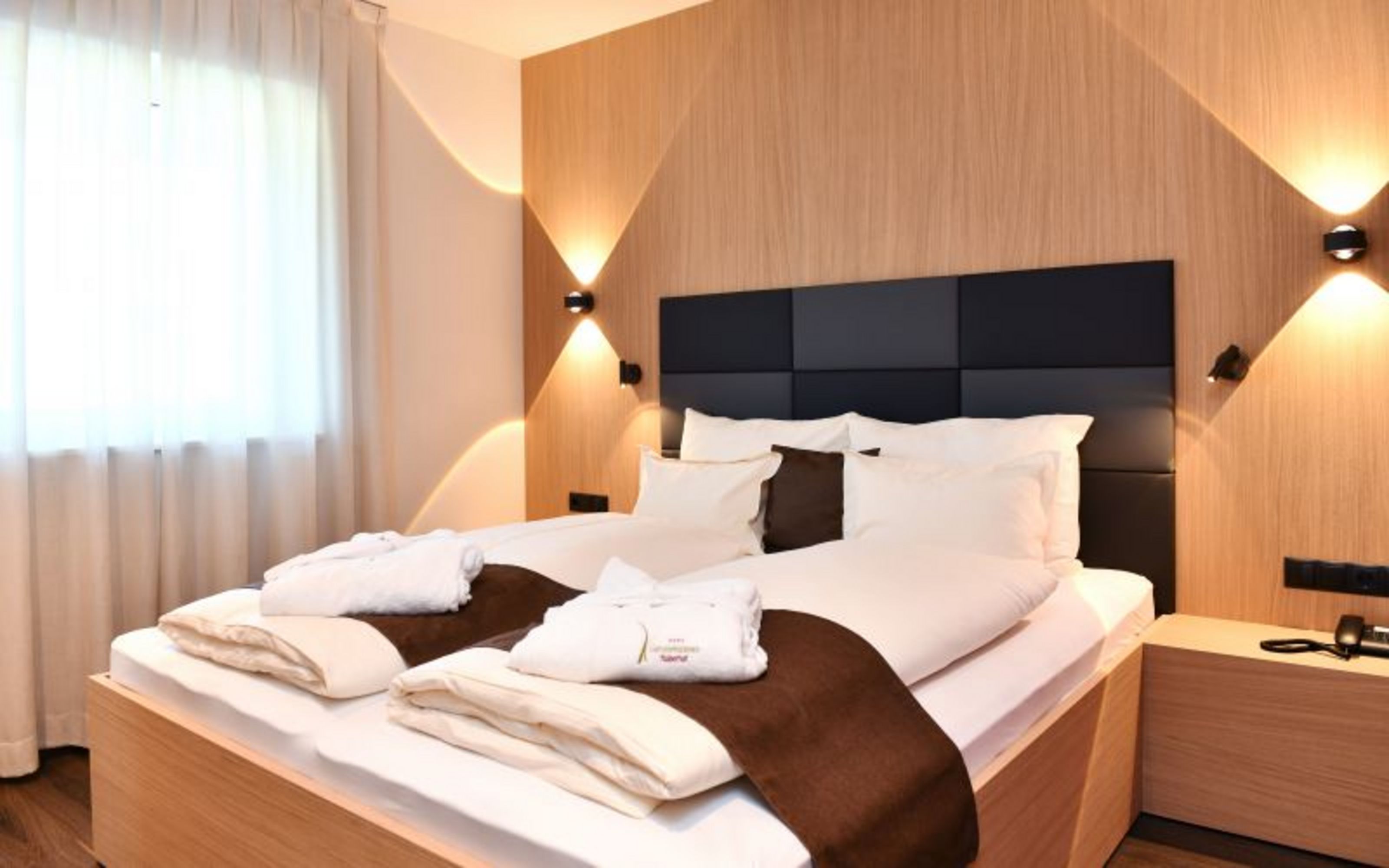Ein modernes Hotelzimmer mit Doppelbett, weißer Bettwäsche, braunen Läufern, Bademänteln und indirekter Beleuchtung vor einer holzverkleideten Wand.