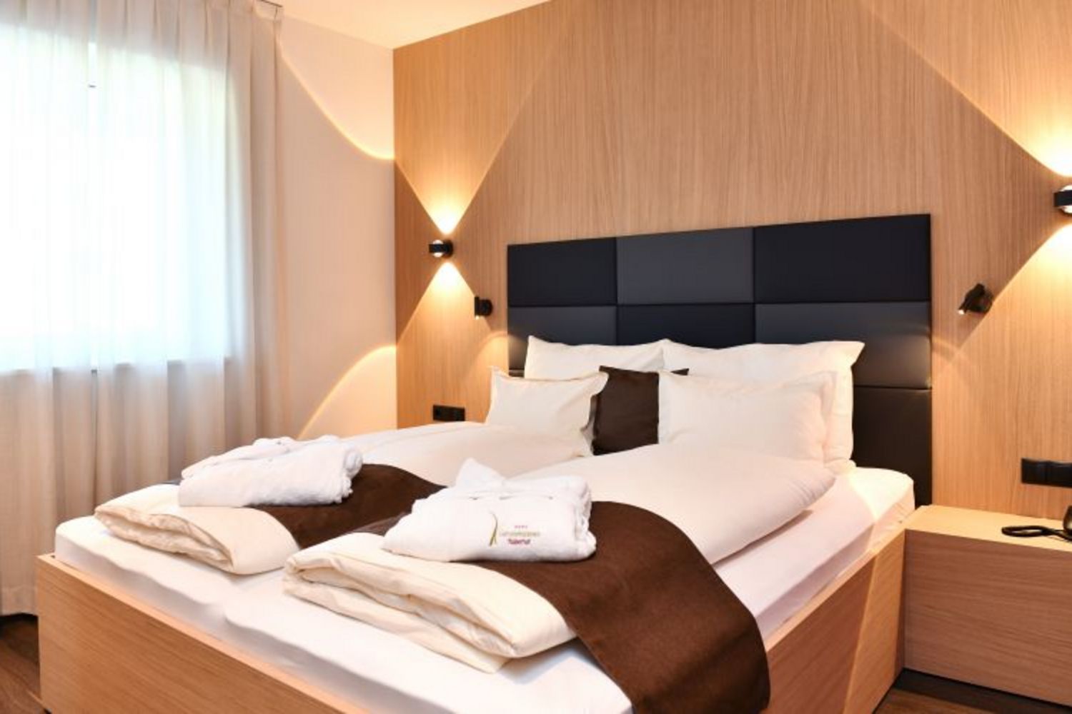Ein modernes Hotelzimmer mit Doppelbett, weißer Bettwäsche, braunen Läufern, Bademänteln und indirekter Beleuchtung vor einer holzverkleideten Wand.