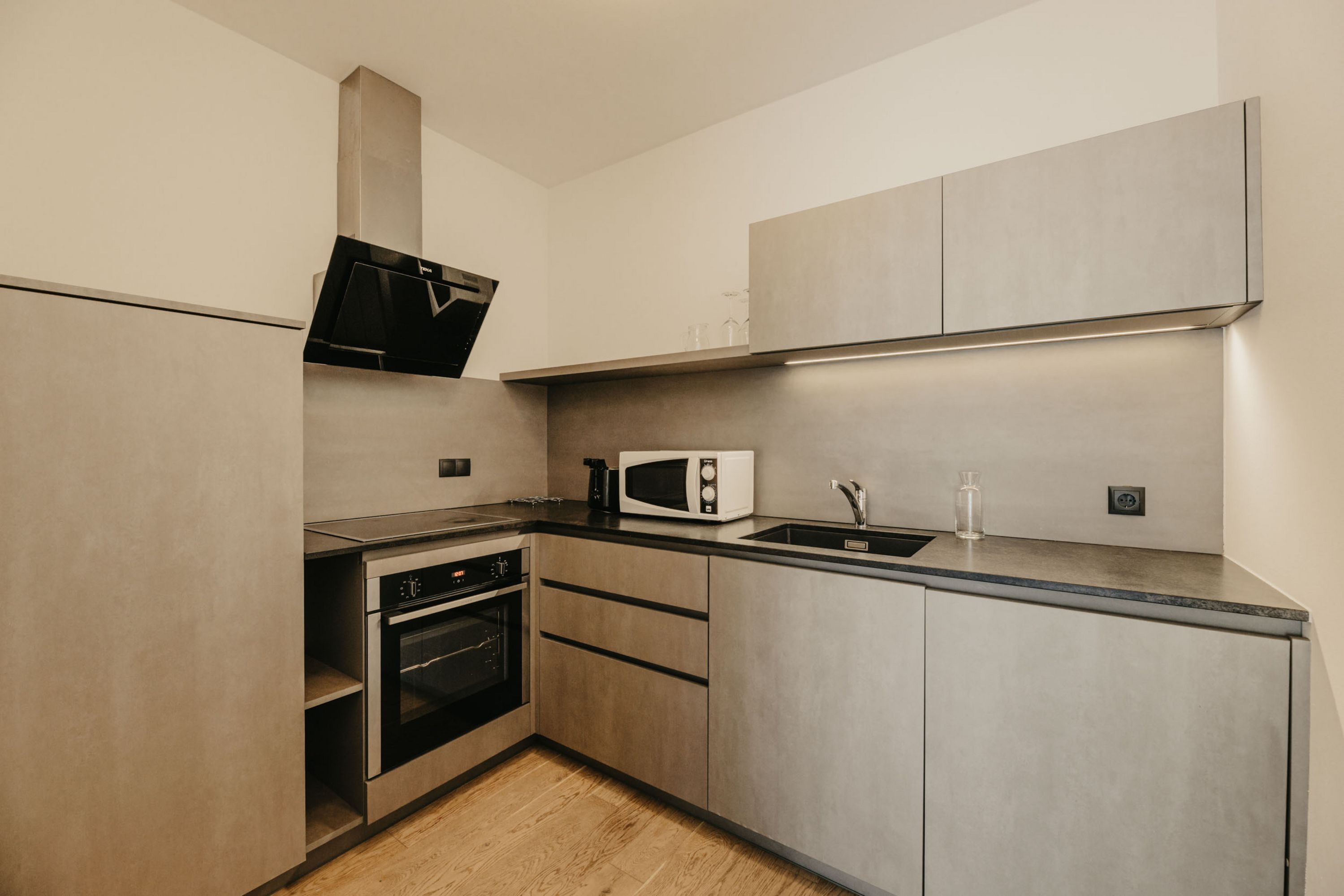 Eine moderne Einbauküche mit grauen Fronten, dunkler Arbeitsplatte, einem Backofen mit Induktionskochfeld, einer Dunstabzugshaube, einer Mikrowelle und einem Spülbecken.
