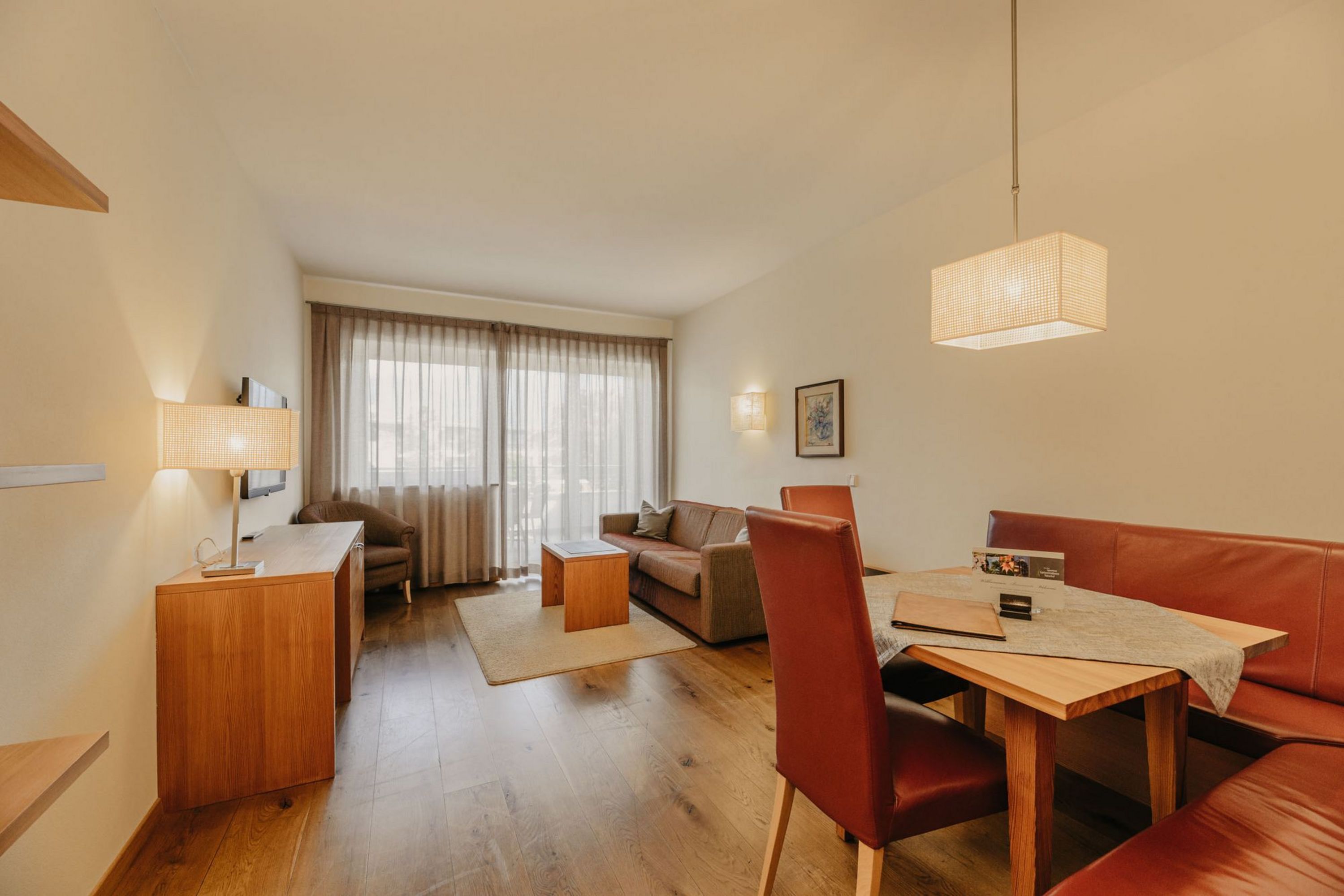 Ein gemütliches Hotelzimmer im modernen Landhausstil mit Holzboden, einer Essecke mit Eckbank und Stühlen sowie einem Wohnbereich mit Sofa und großen Fenstern.
