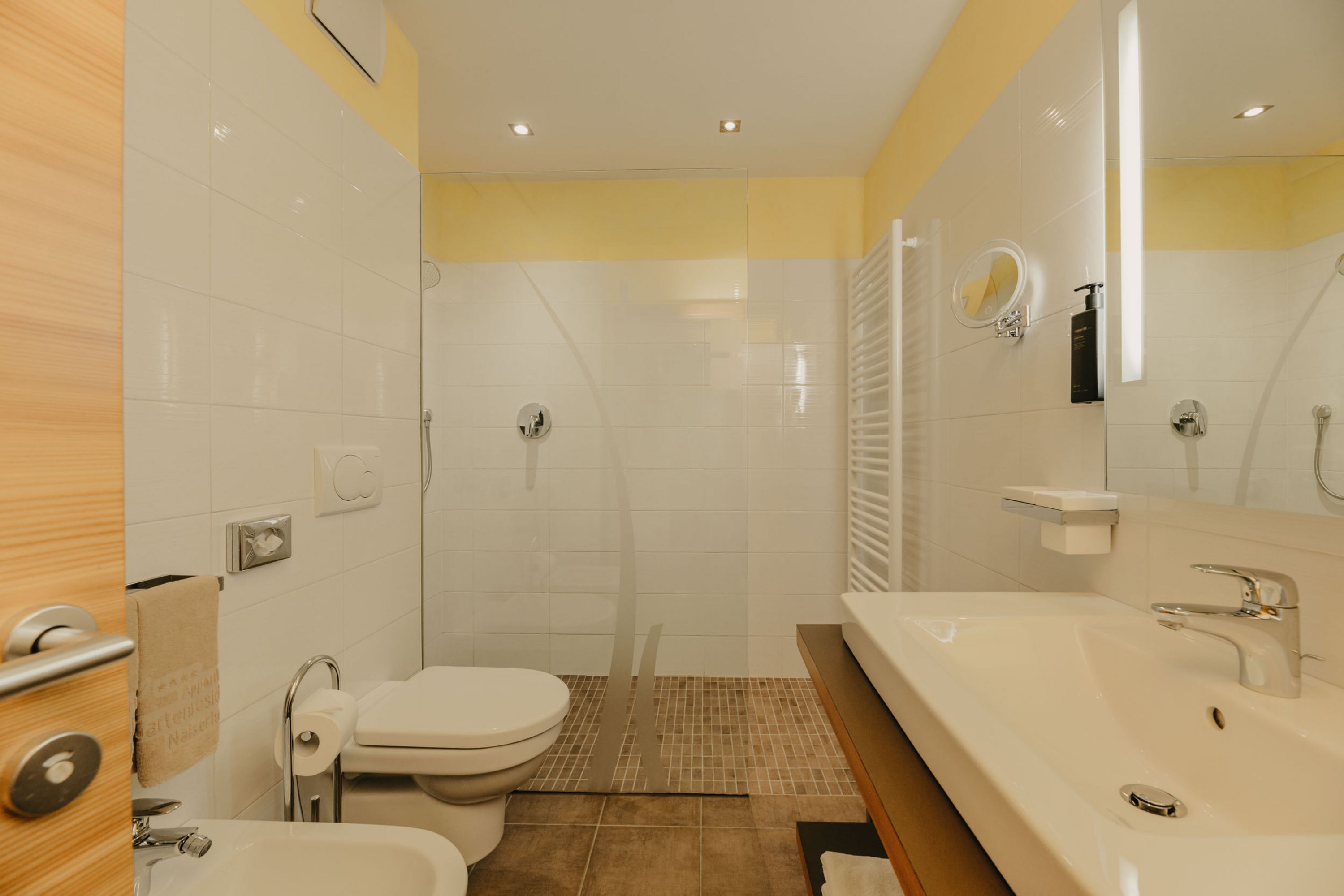 Ein modernes Badezimmer mit weiß gefliesten Wänden, einer offenen bodengleichen Dusche mit Glaswand, einem Waschbecken und einem WC.