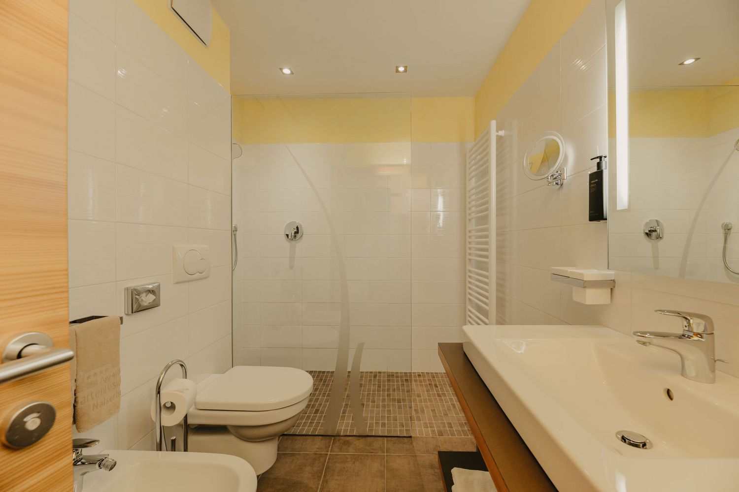 Ein modernes Badezimmer mit weiß gefliesten Wänden, einer offenen bodengleichen Dusche mit Glaswand, einem Waschbecken und einem WC.