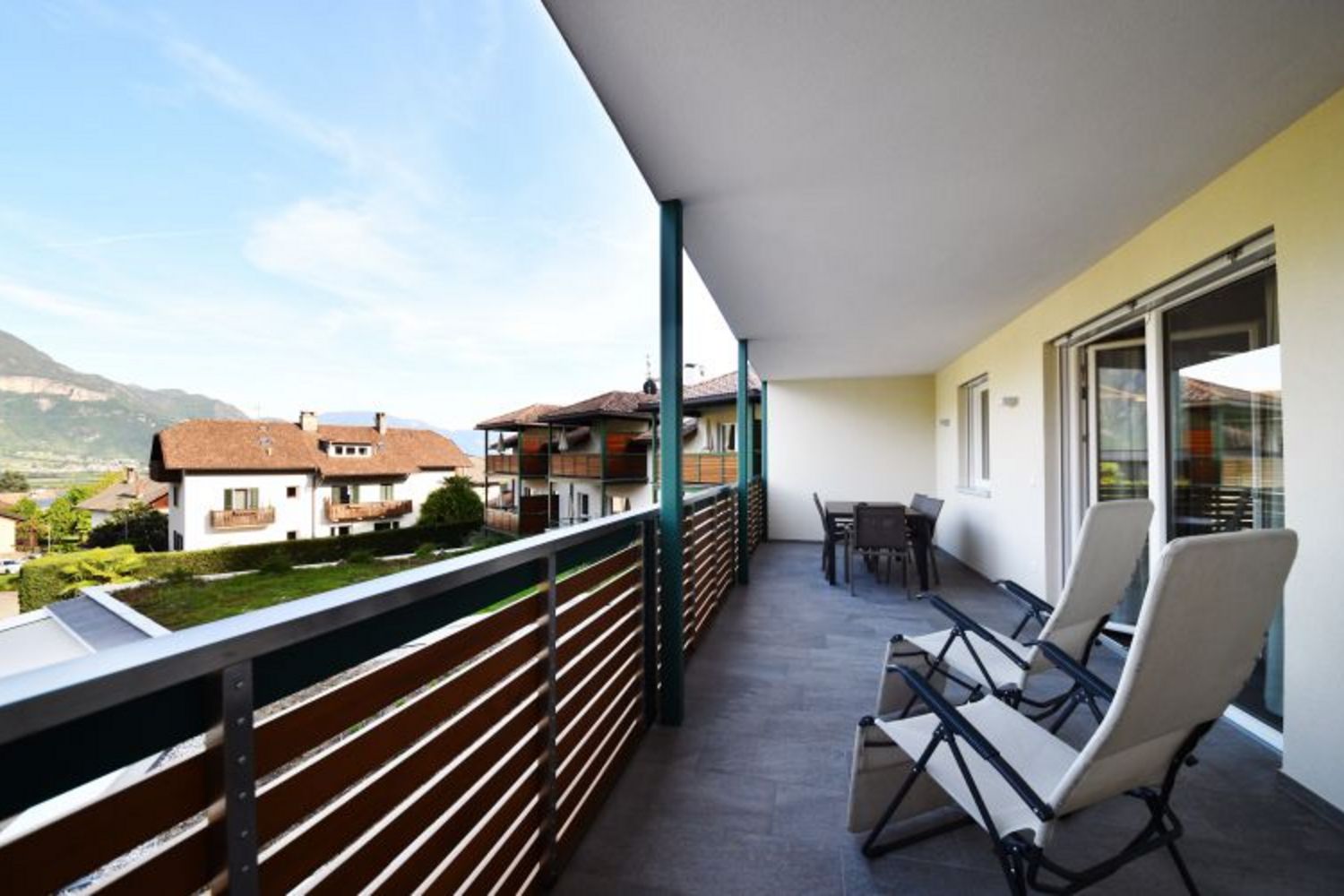 Ein weitläufiger Balkon mit Fliesenboden, zwei Liegestühlen, einem kleinen Tisch mit Stühlen und Blick auf umliegende Häuser und Berge.