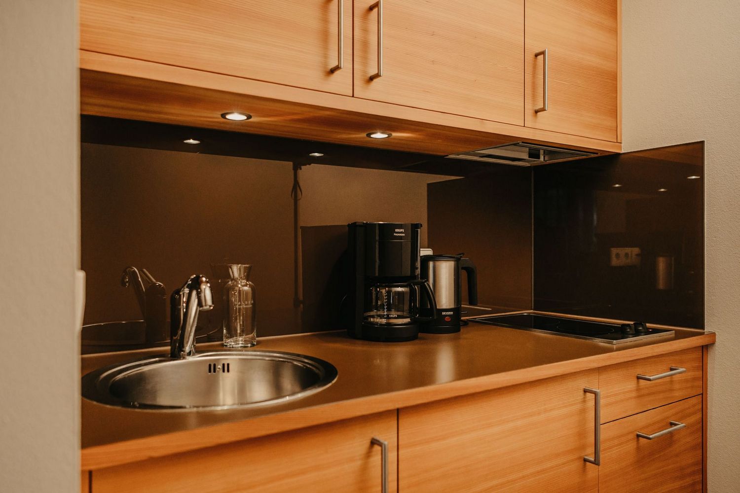 Eine moderne Küchenzeile mit Holzschränken, Spüle, Kaffeemaschine und Herdplatte vor einer dunklen Glasrückwand.