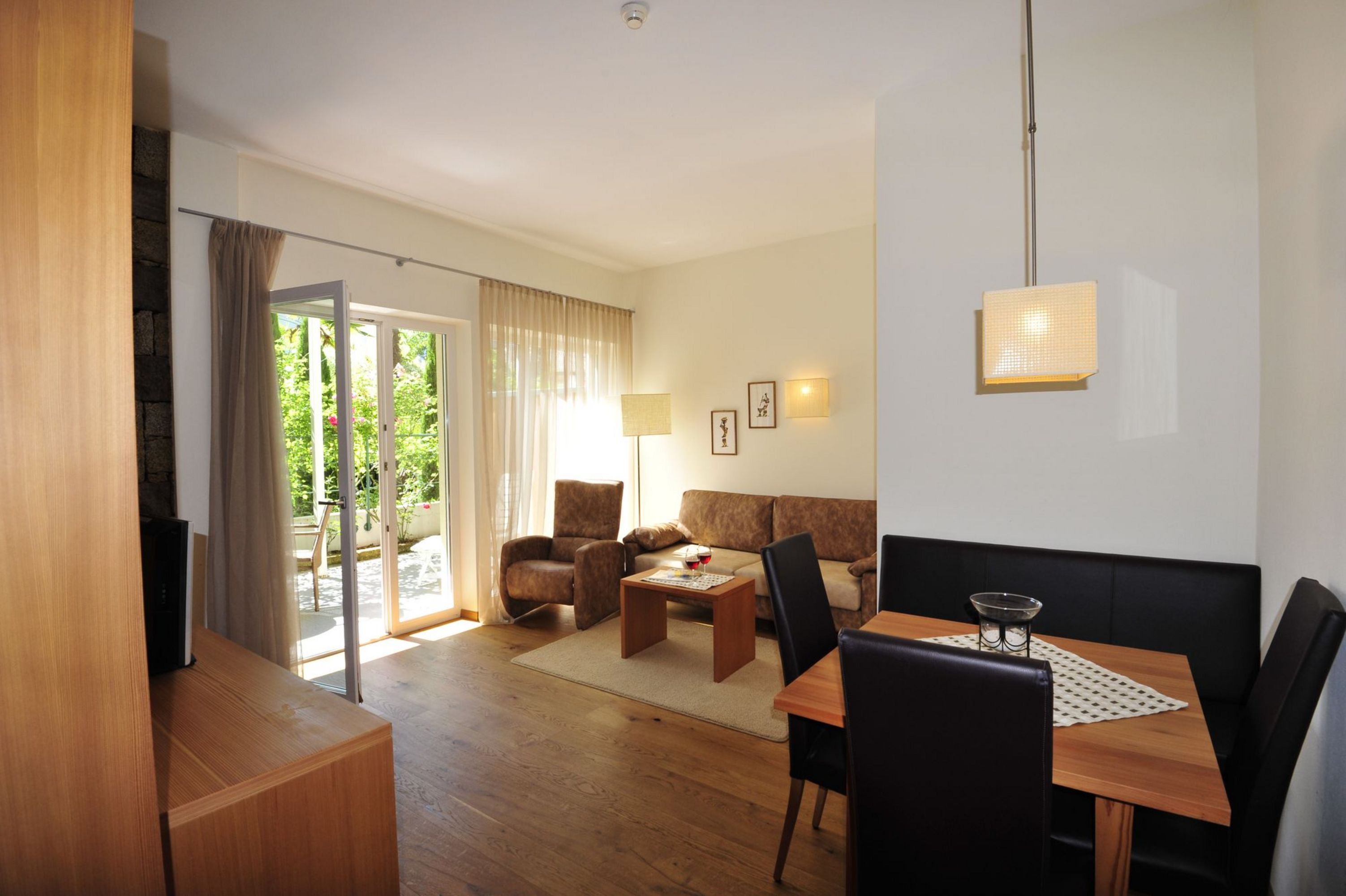 Ein lichtdurchfluteter Wohnraum mit einer Sitzecke aus Sofa und Sessel, einem kleinen Esstisch mit schwarzen Stühlen und einer Terrassentür, die nach draußen führt.