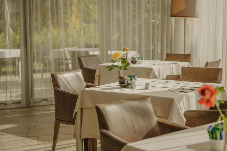 Ein helles Restaurant-Interieur mit gedeckten Tischen, braunen Lederstühlen und einer einzelnen gelben Rose in einer Vase auf dem mittleren Tisch.