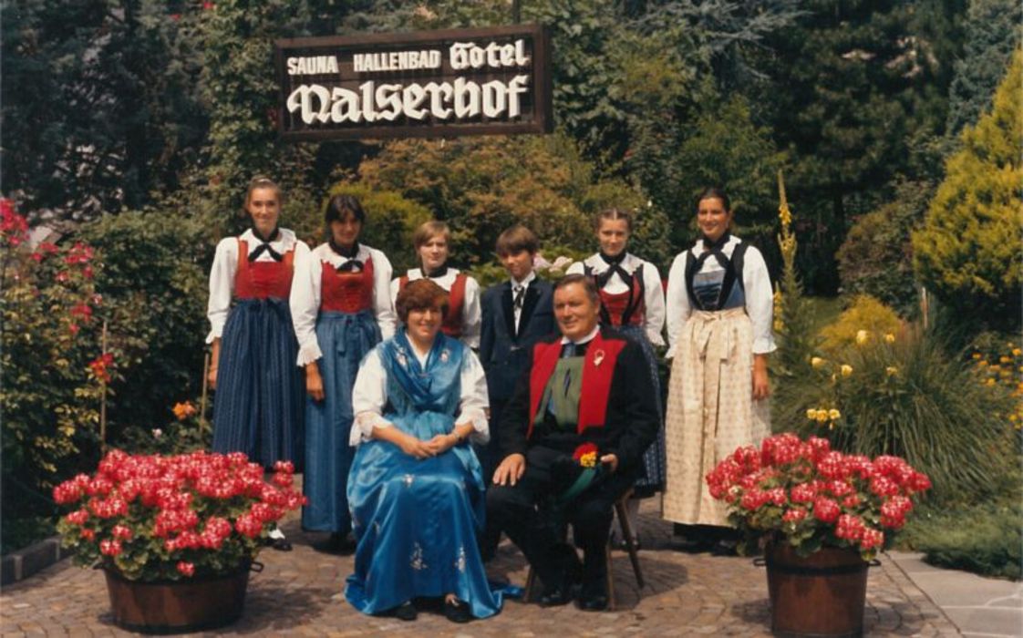 Eine Gruppe von zwei Erwachsenen und vier Jugendlichen in traditioneller Südtiroler Tracht vor einem Hotel-Schild mit der Aufschrift Nalserhof.
