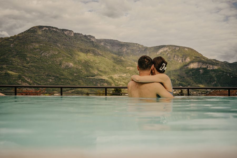 Ein junges Paar umarmt sich von hinten im Wasser eines Infinity-Pools vor einer bergigen Landschaft.