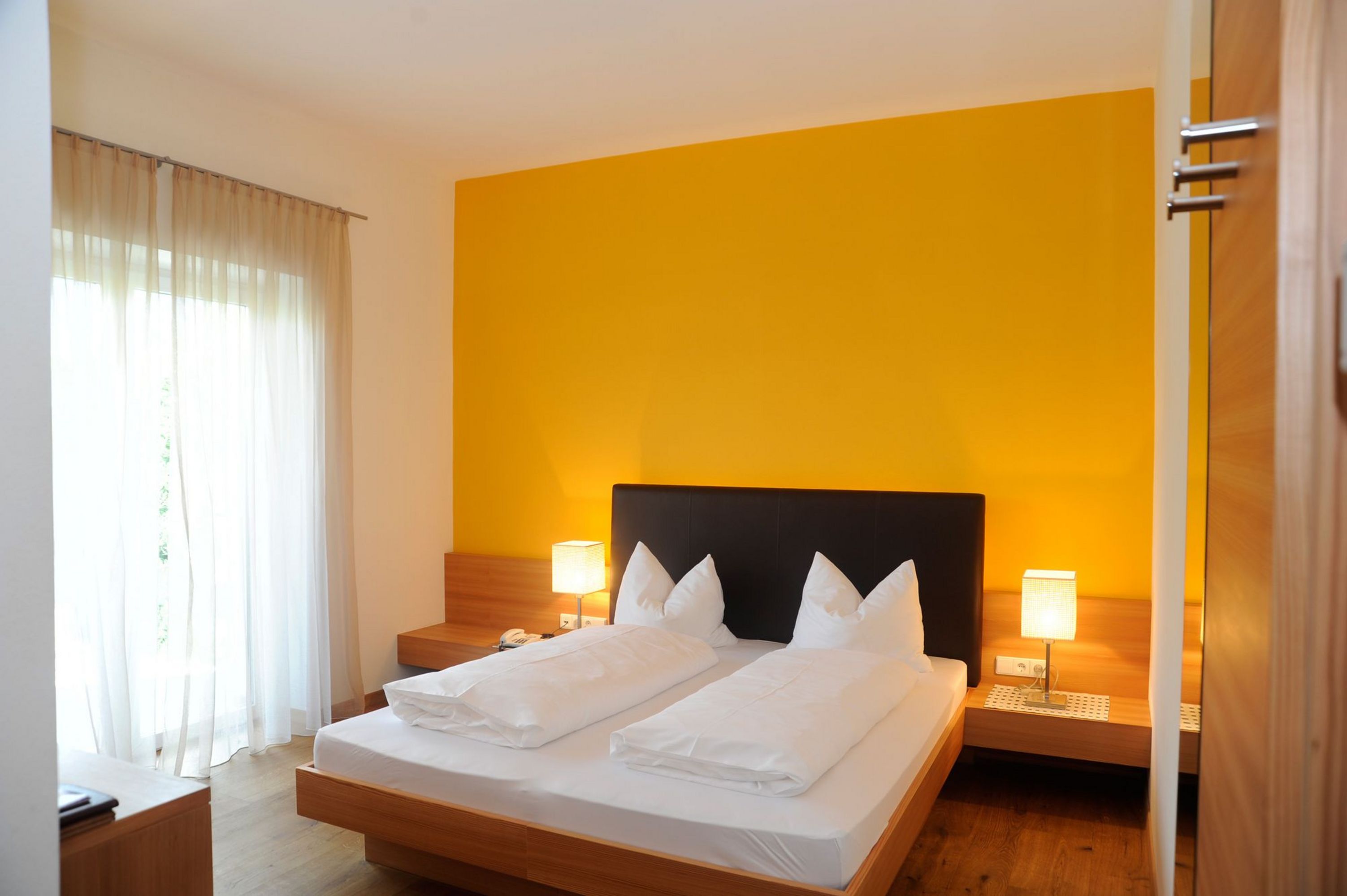 Ein Hotelzimmer mit einem Doppelbett, einem dunklen Kopfteil, zwei Nachttischlampen und einer markanten, sonnengelben Wand hinter dem Bett.