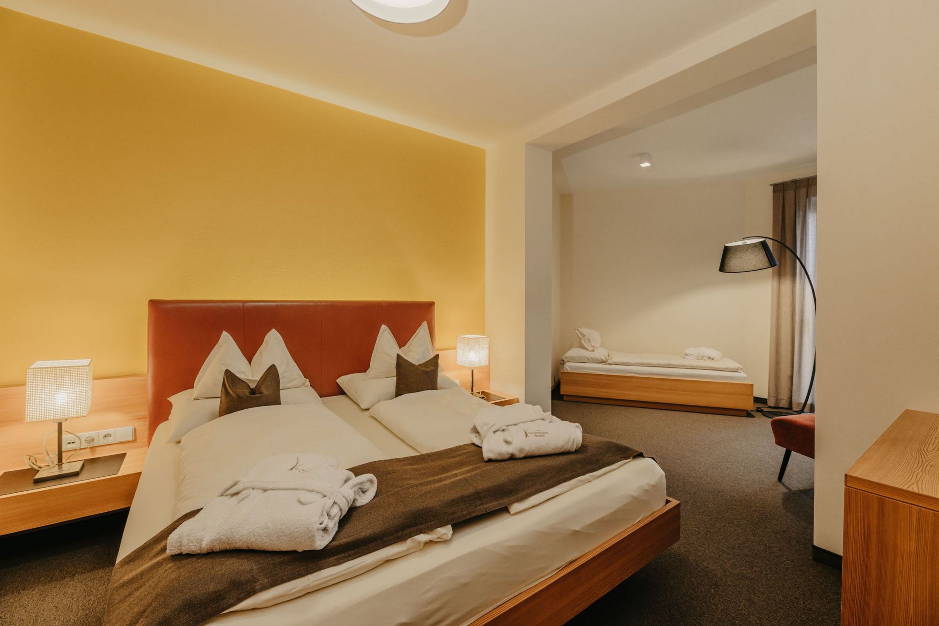 Ein Hotelzimmer mit einem Doppelbett, zwei Bademänteln darauf und einem zusätzlichen Einzelbett im angrenzenden Wohnbereich.