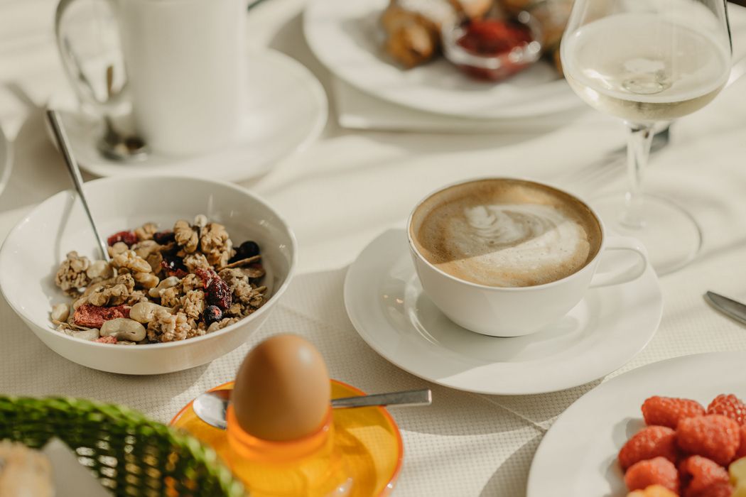 Ein reich gedeckter Frühstückstisch mit einer Schale Müsli, einem Cappuccino, einem Frühstücksei und einem Teller mit frischen Himbeeren im Sonnenlicht.