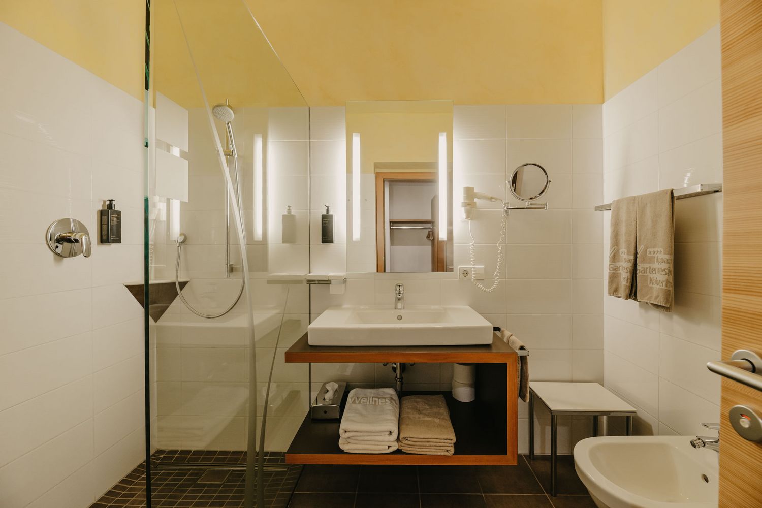 Ein modernes Badezimmer mit einer bodengleichen Glasdusche, einem Waschbecken auf einem Holzwaschtisch mit Handtuchablage und einem Bidet.