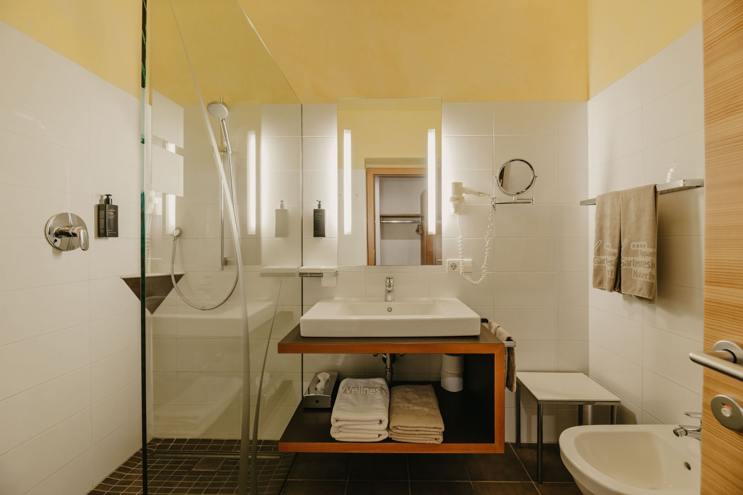 Ein modernes Badezimmer mit einer bodengleichen Glasdusche, einem Waschbecken auf einem Holzwaschtisch mit Handtuchablage und einem Bidet.