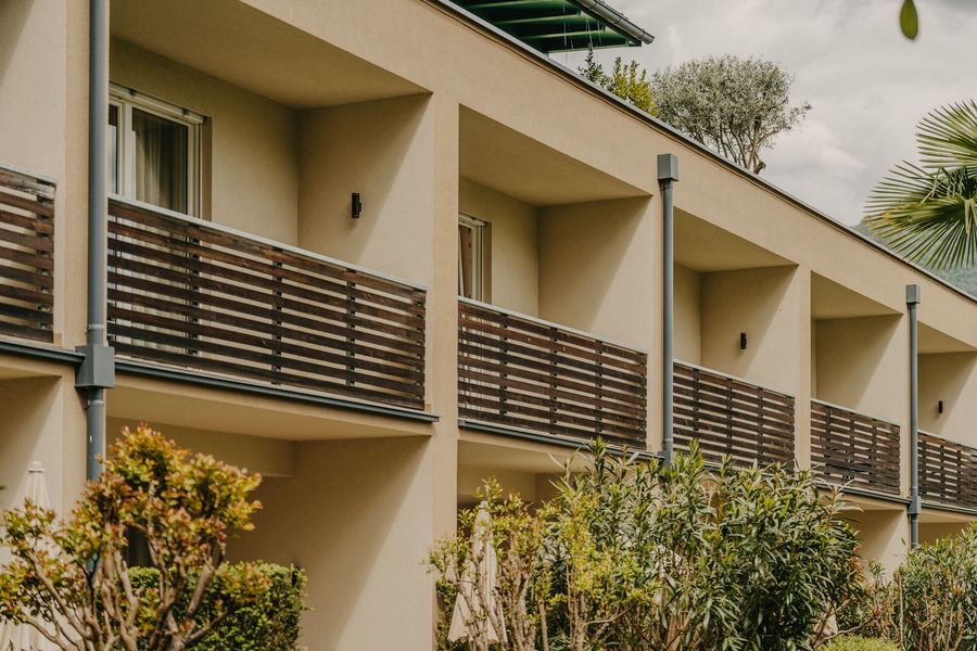 Eine Fassade eines modernen Hotelgebäudes mit Balkonen, die aus beigen Wänden und horizontalen Holzelementen bestehen, umrahmt von mediterraner Vegetation.