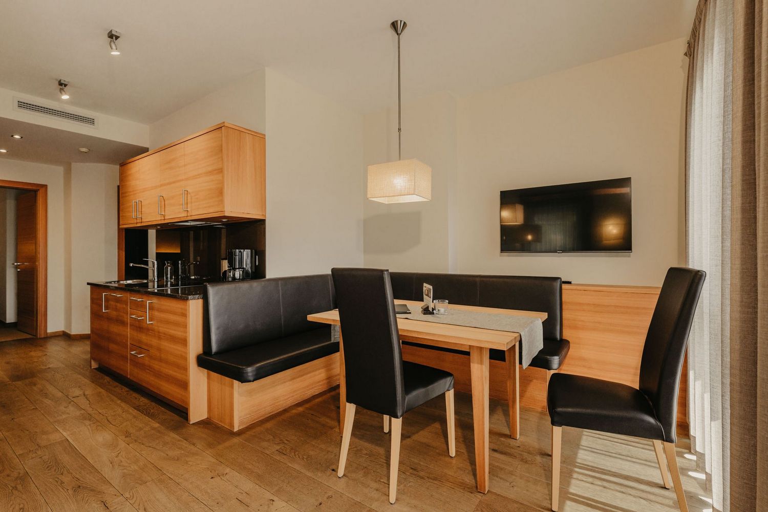 Ein offener Wohnbereich mit einer modernen Einbauküche aus Holz und einem angrenzenden Essbereich mit einer schwarzen Sitzbank und Stühlen an einem Holztisch.
