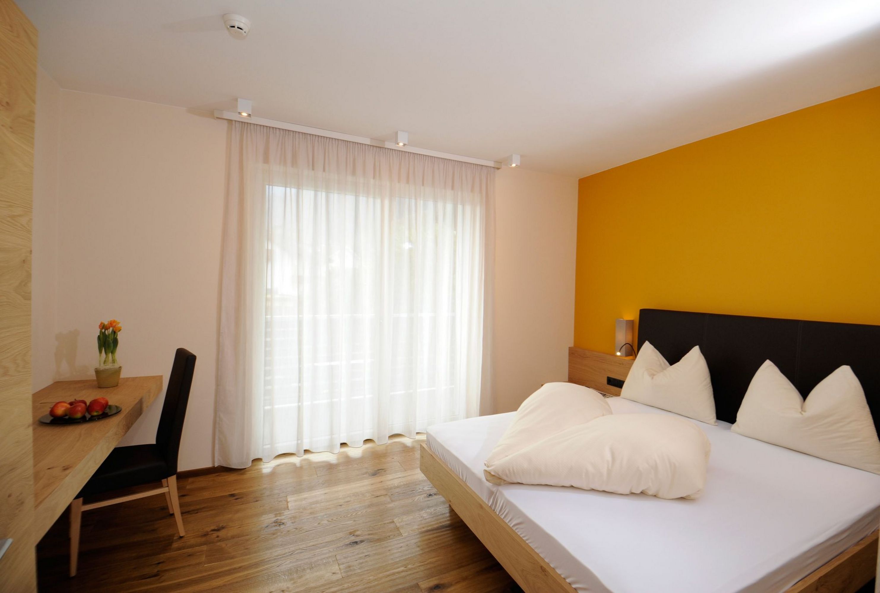 Ein helles Hotelzimmer mit gelber Akzentwand, Holzboden, einem Doppelbett mit zwei Kissen und einer herzförmigen Decke, sowie einem Schreibtisch mit Stuhl und Äpfeln.