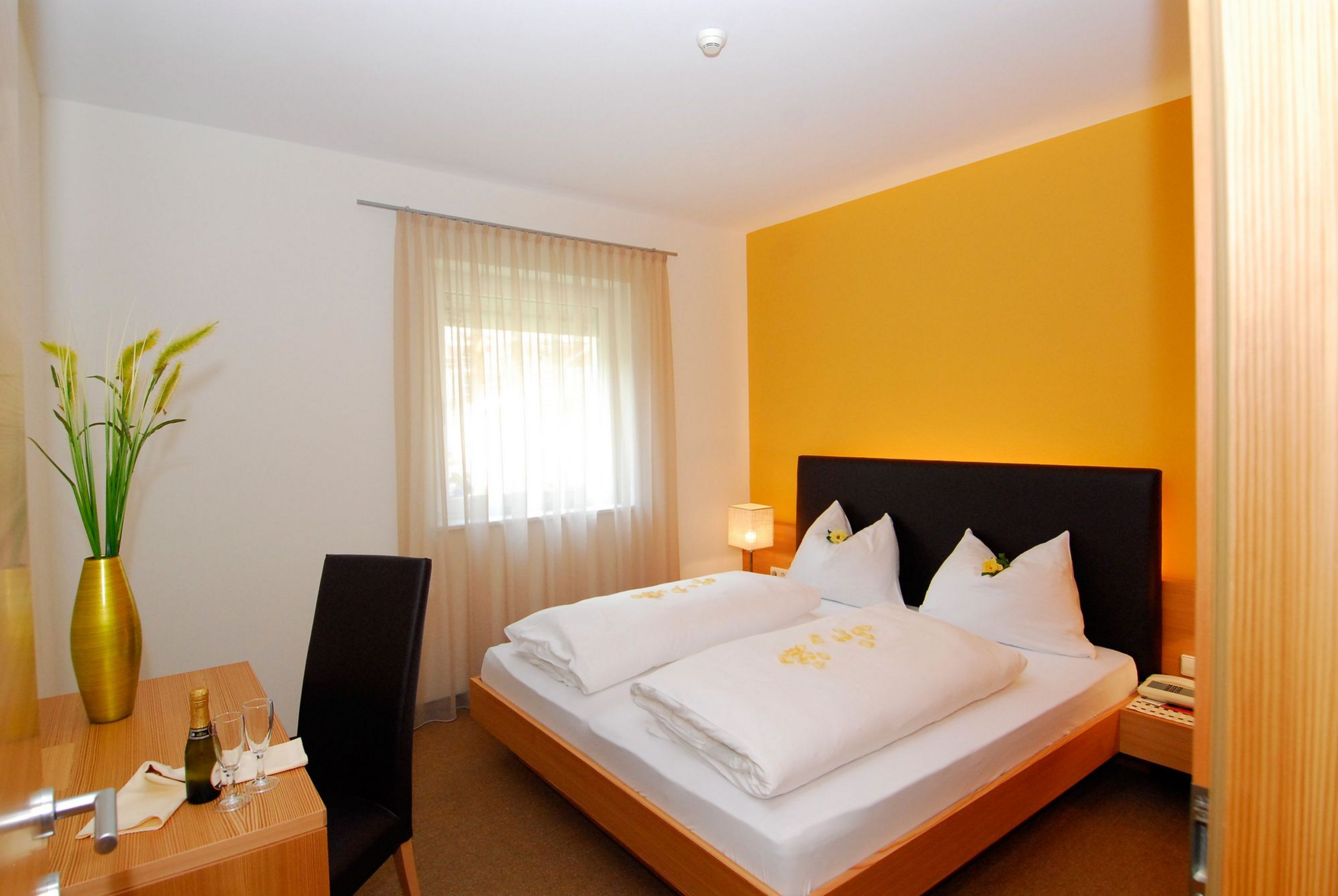 Ein einladendes Hotelzimmer mit einem Doppelbett, gelber Akzentwand und einem Schreibtisch mit Sektflasche.