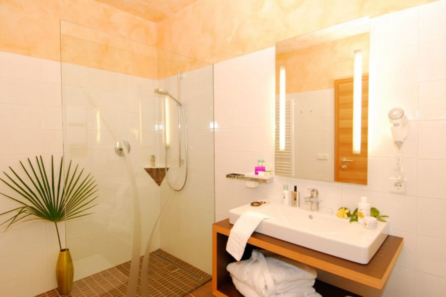 Ein helles, modernes Hotelbadezimmer mit einer gläsernen Walk-in-Dusche, einem Waschbecken auf einem Holzunterschrank, einem beleuchteten Spiegel und einem Föhn an der Wand.