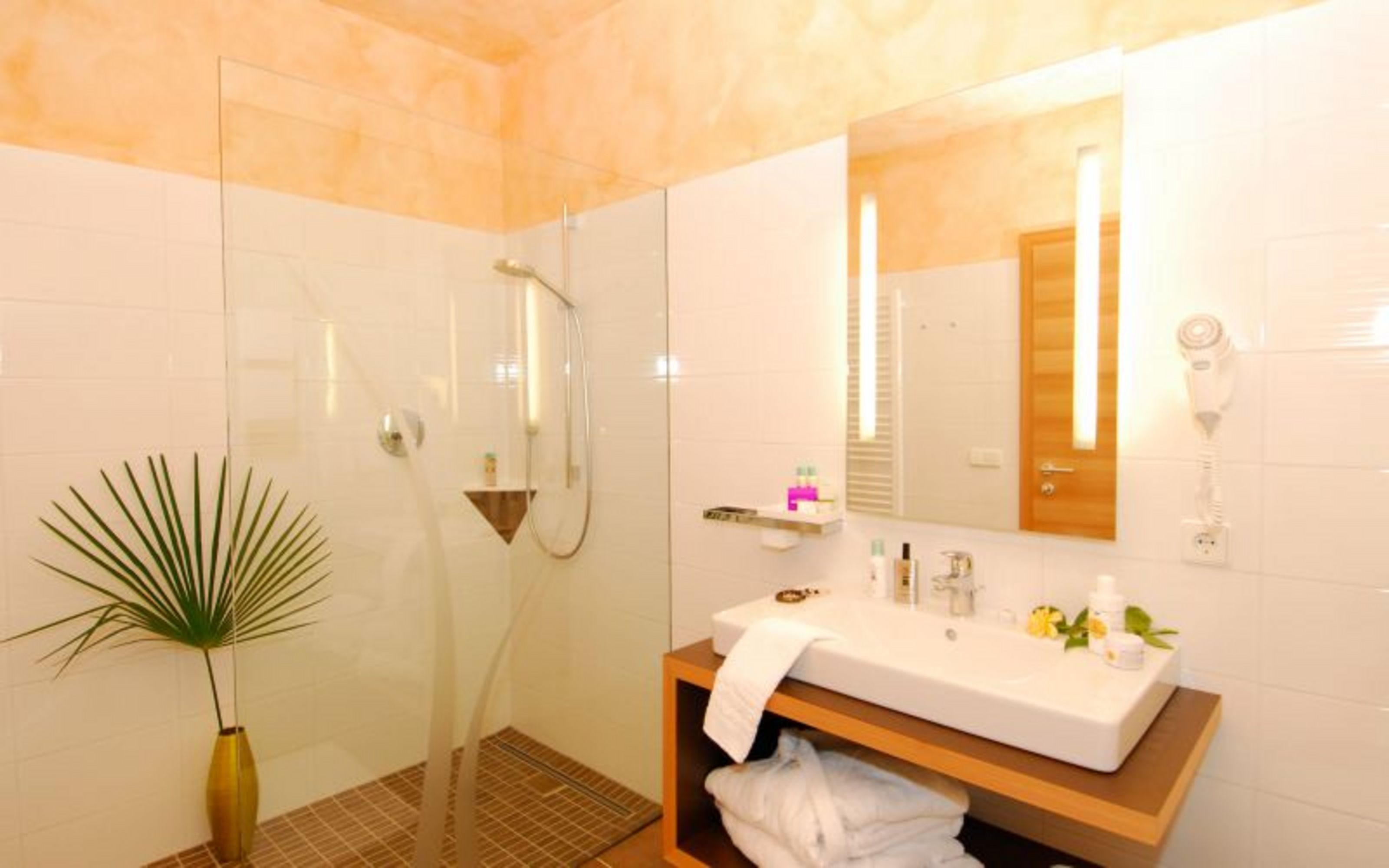 Ein helles, modernes Hotelbadezimmer mit einer gläsernen Walk-in-Dusche, einem Waschbecken auf einem Holzunterschrank, einem beleuchteten Spiegel und einem Föhn an der Wand.