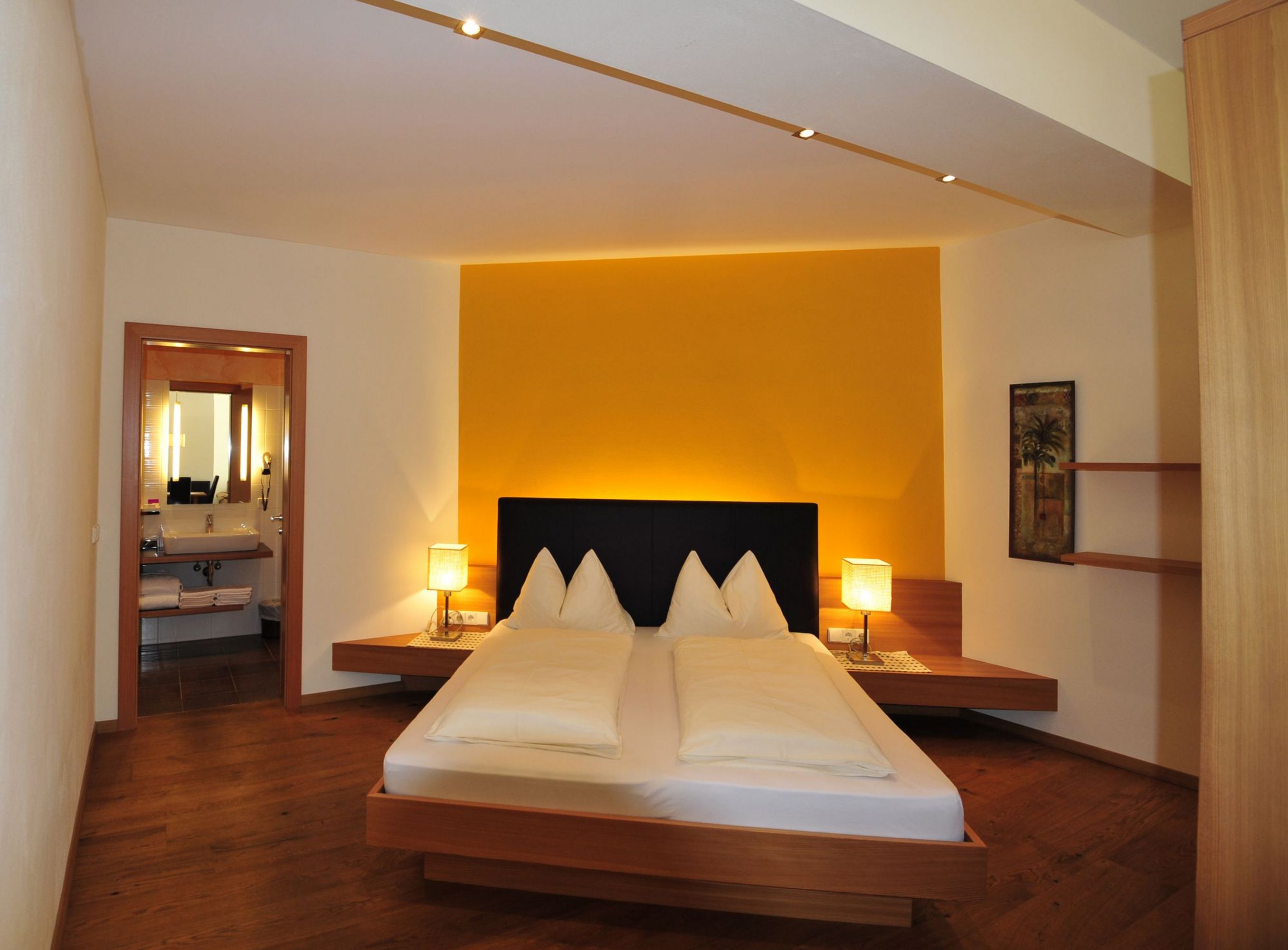 Ein modernes Hotelzimmer mit Doppelbett, leuchtend gelber Akzentwand hinter dem Bett, schwebenden Nachttischen mit Lampen und Blick in ein angrenzendes Badezimmer.