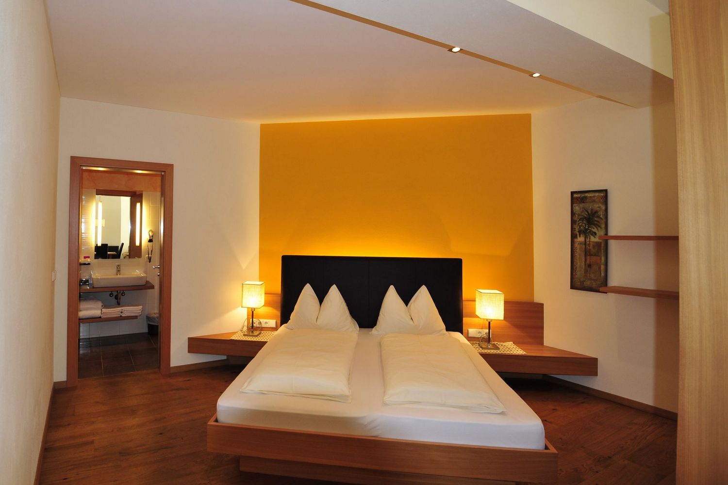 Ein modernes Hotelzimmer mit Doppelbett, leuchtend gelber Akzentwand hinter dem Bett, schwebenden Nachttischen mit Lampen und Blick in ein angrenzendes Badezimmer.