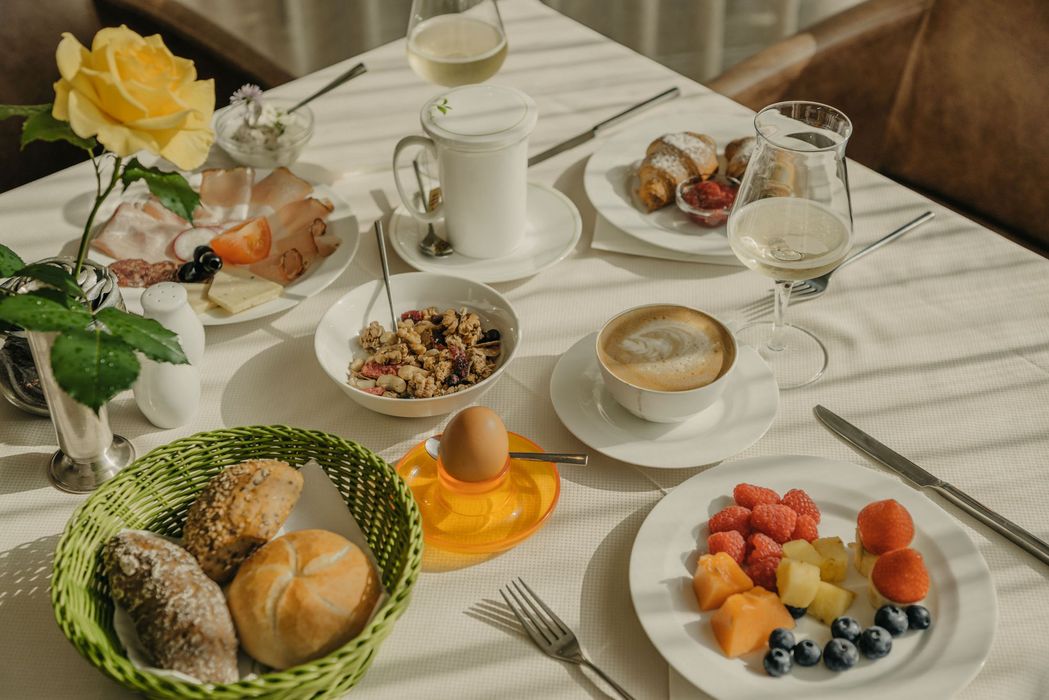 Ein reichhaltig gedeckter Frühstückstisch mit Brötchenkorb, Müsli, Aufschnitt, Obstteller, gekochtem Ei, Kaffee und einem Glas Weißwein.